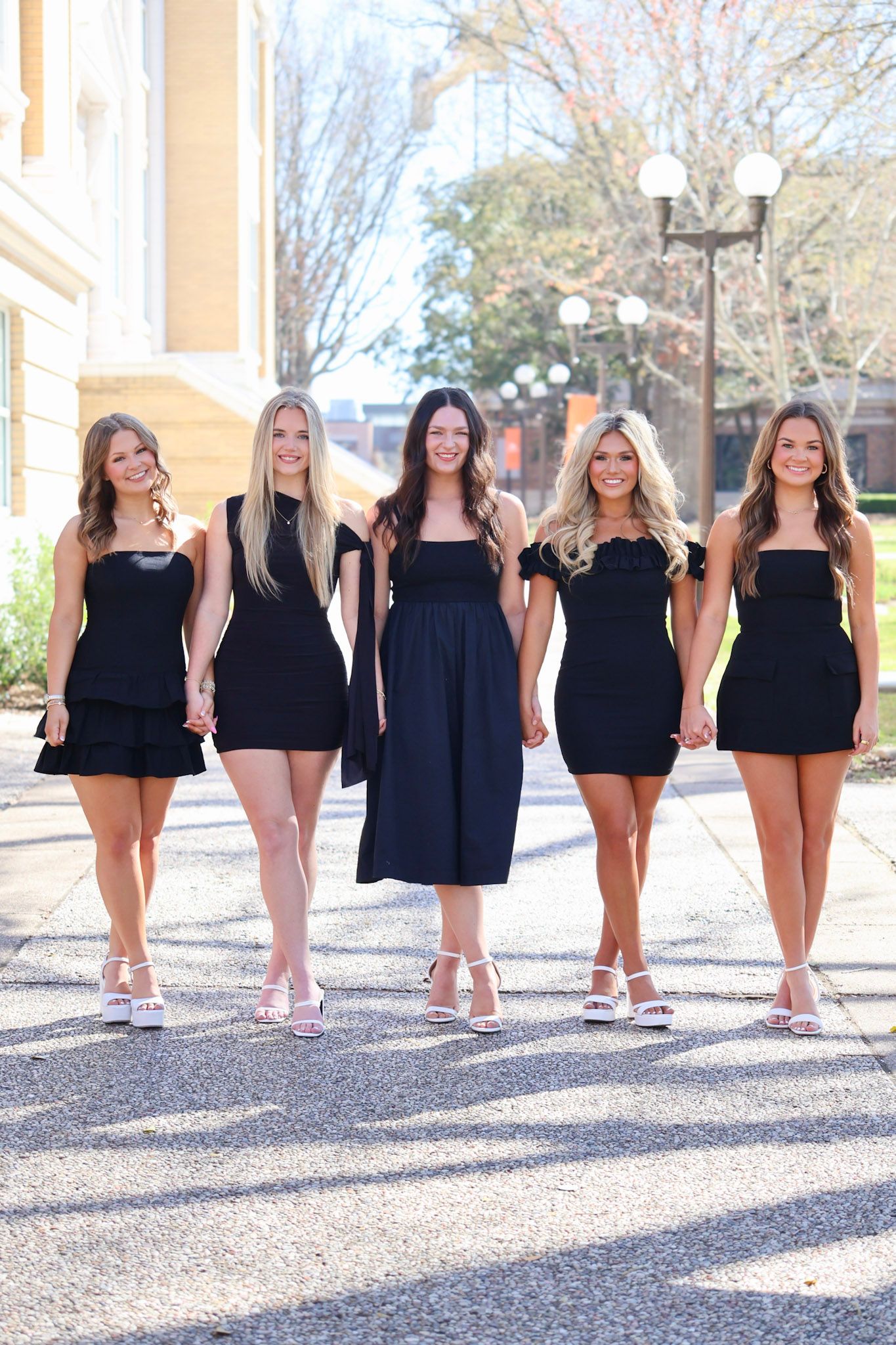 Five young women in matching black dresses and white heels walking together outdoors near a classic building.