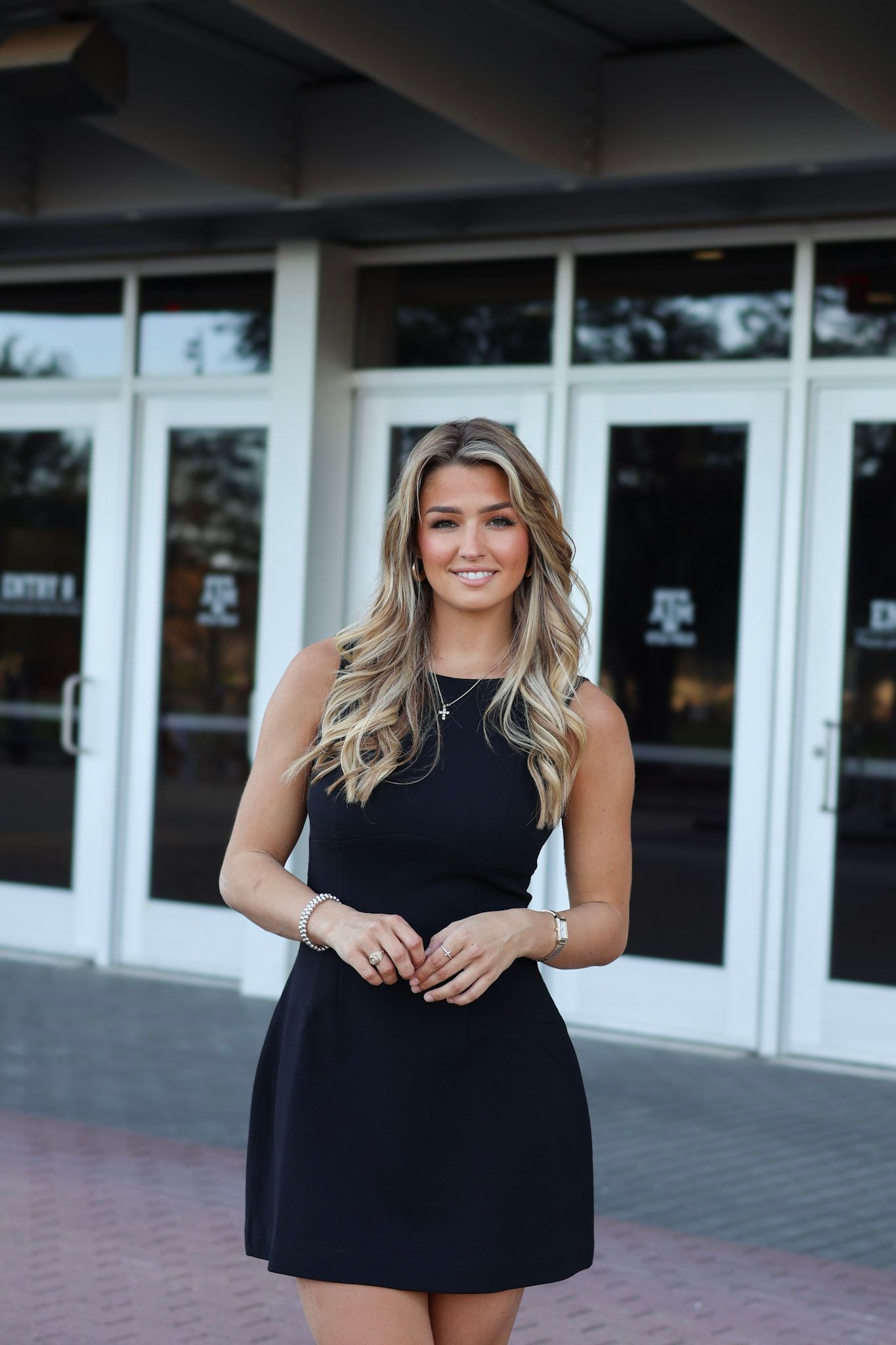 Young woman in black sleeveless dress posing outdoors near glass doors of a modern building.