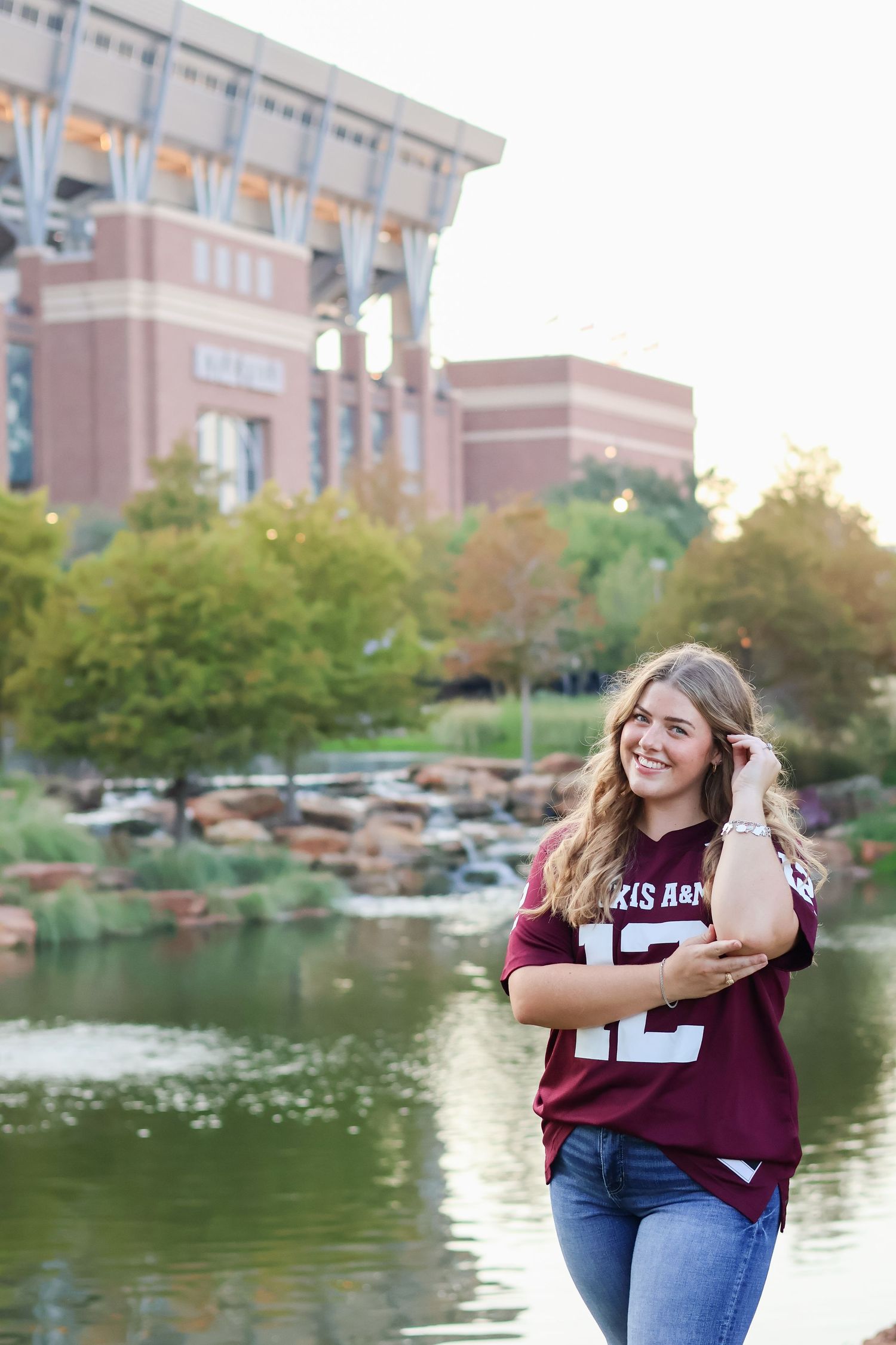 A student in a maroon Texas A&M jersey poses by a scenic campus pond with stadium architecture in the background.