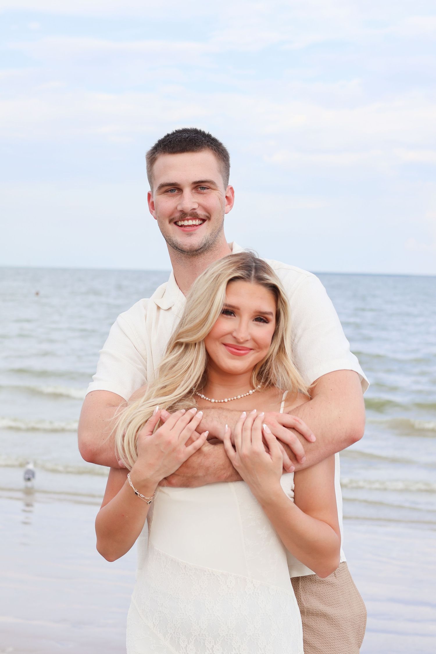 A couple embraces joyfully on a sunny beach with waves and sand in the background.
