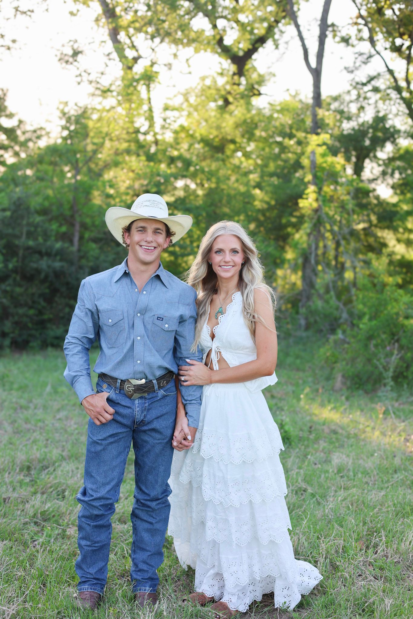 A smiling couple in a field, man in cowboy hat and jeans, woman in white lace dress, posing together outdoors.