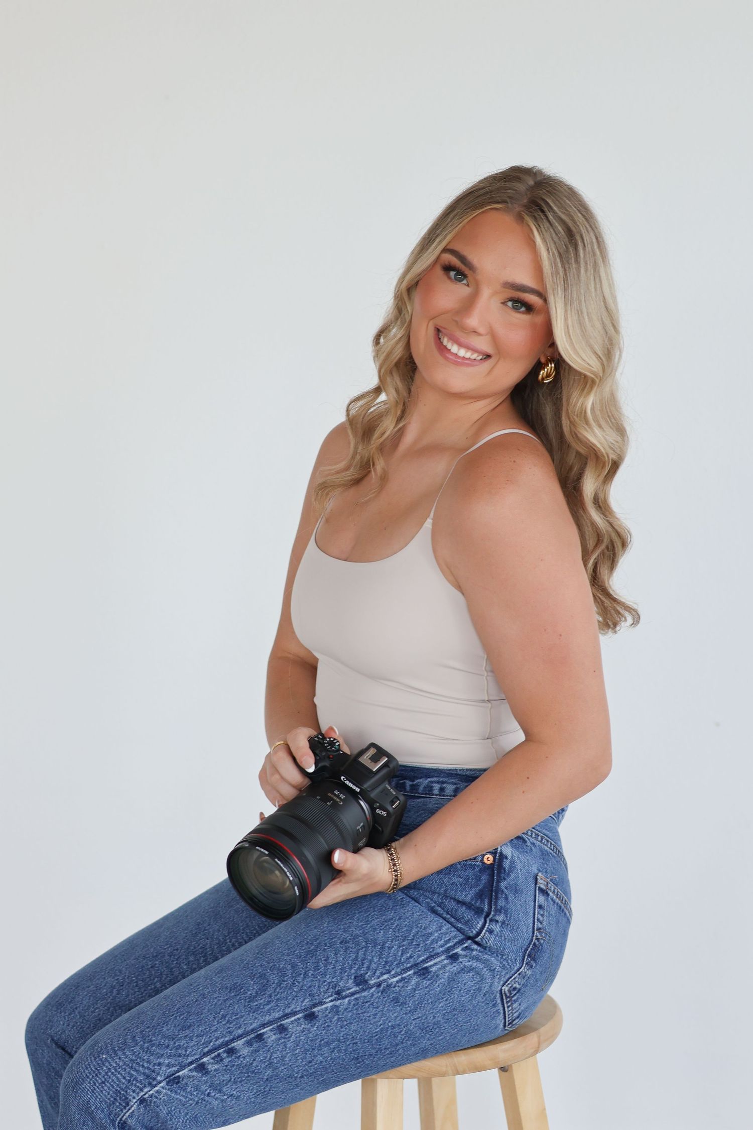 Professional photographer in casual attire poses with camera on wooden stool against white studio backdrop.