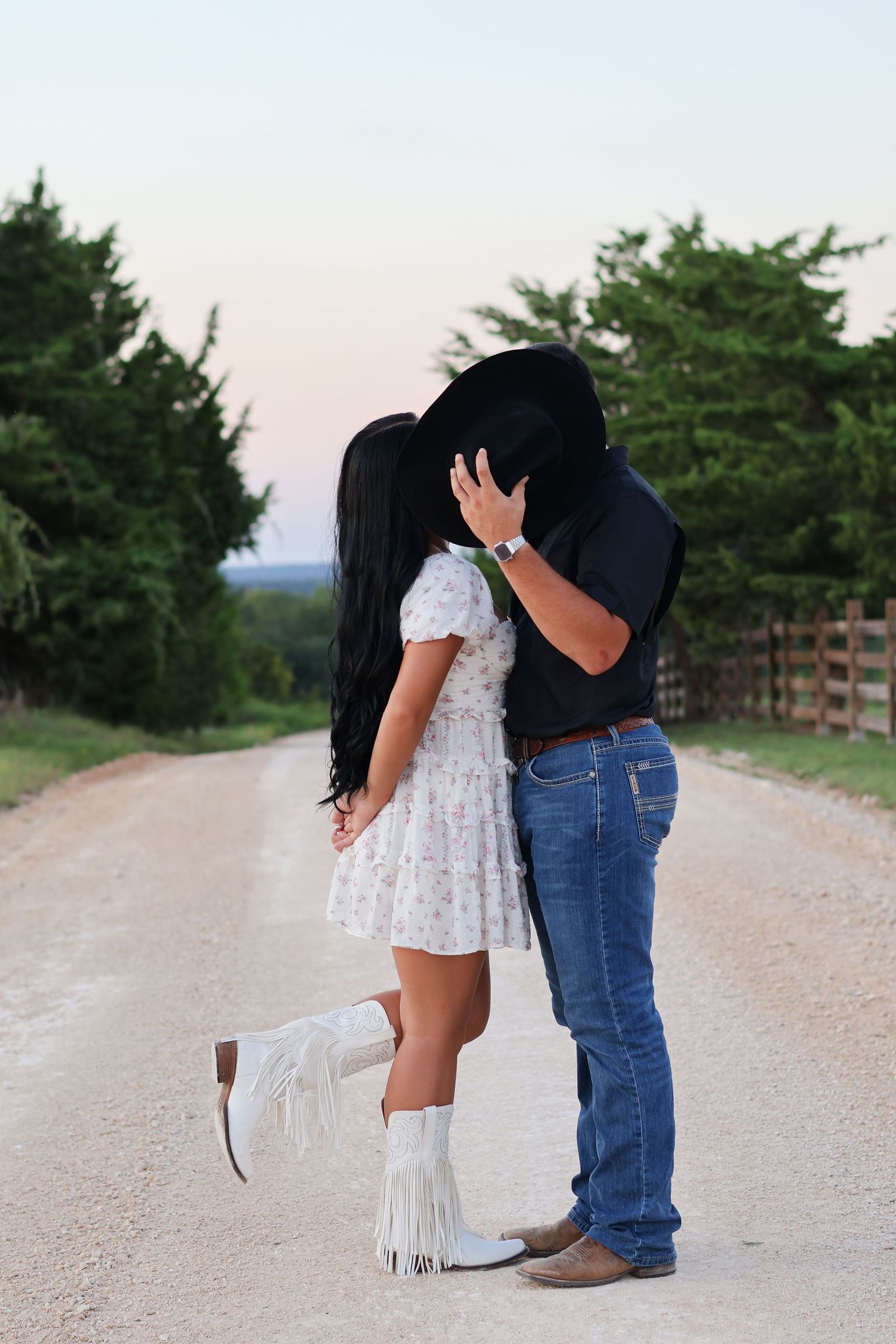 A couple in cowboy boots embrace on a dirt road surrounded by green trees and countryside scenery.