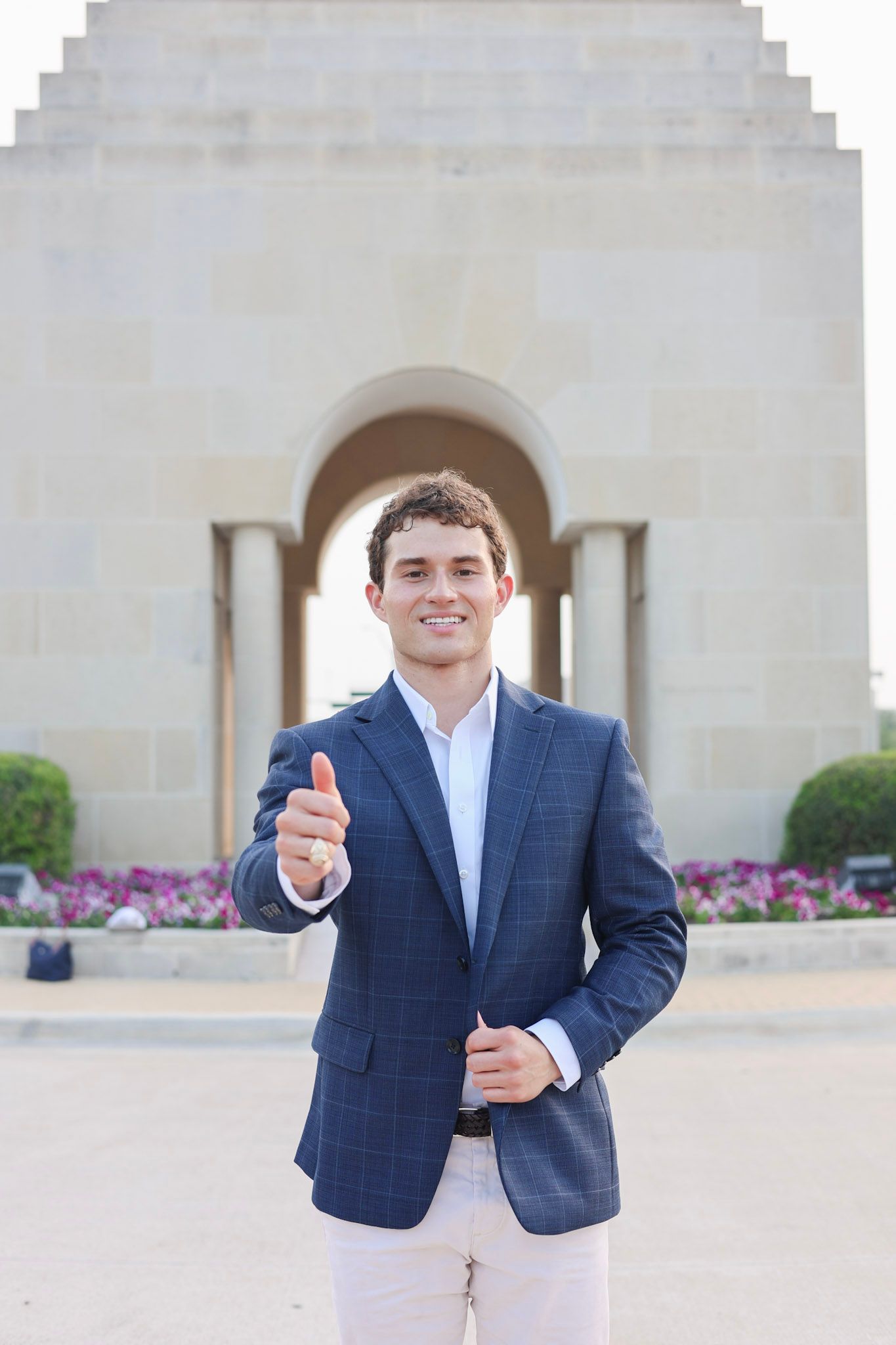 Young man in blue blazer giving thumbs up in front of a stone archway monument with flowers.