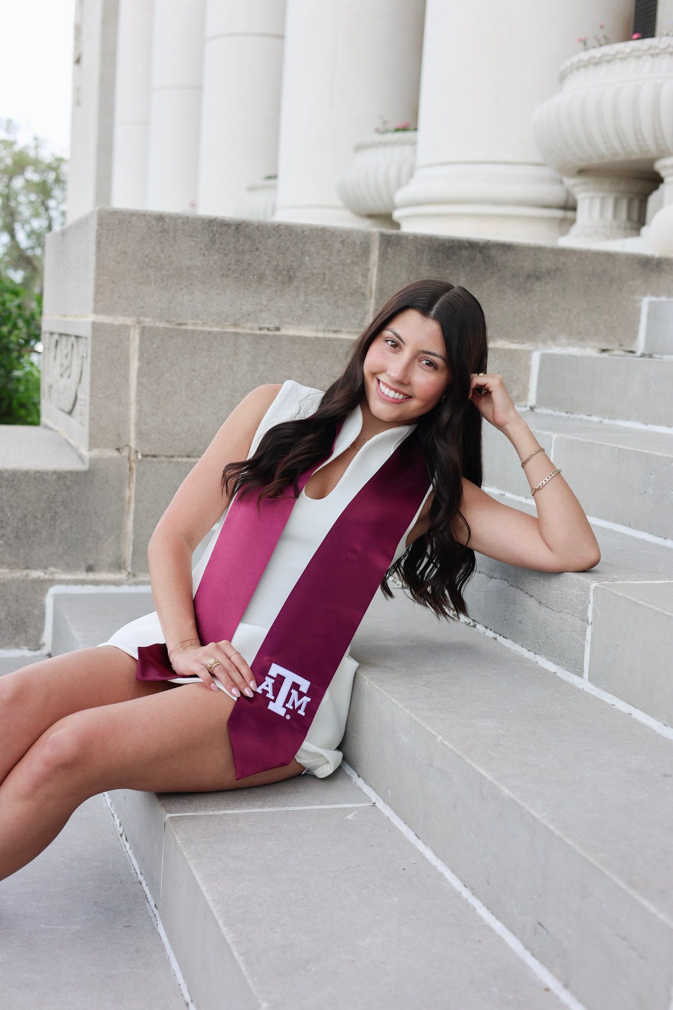 Smiling young woman in white dress wearing Texas A&M maroon graduation stole seated on stone steps.