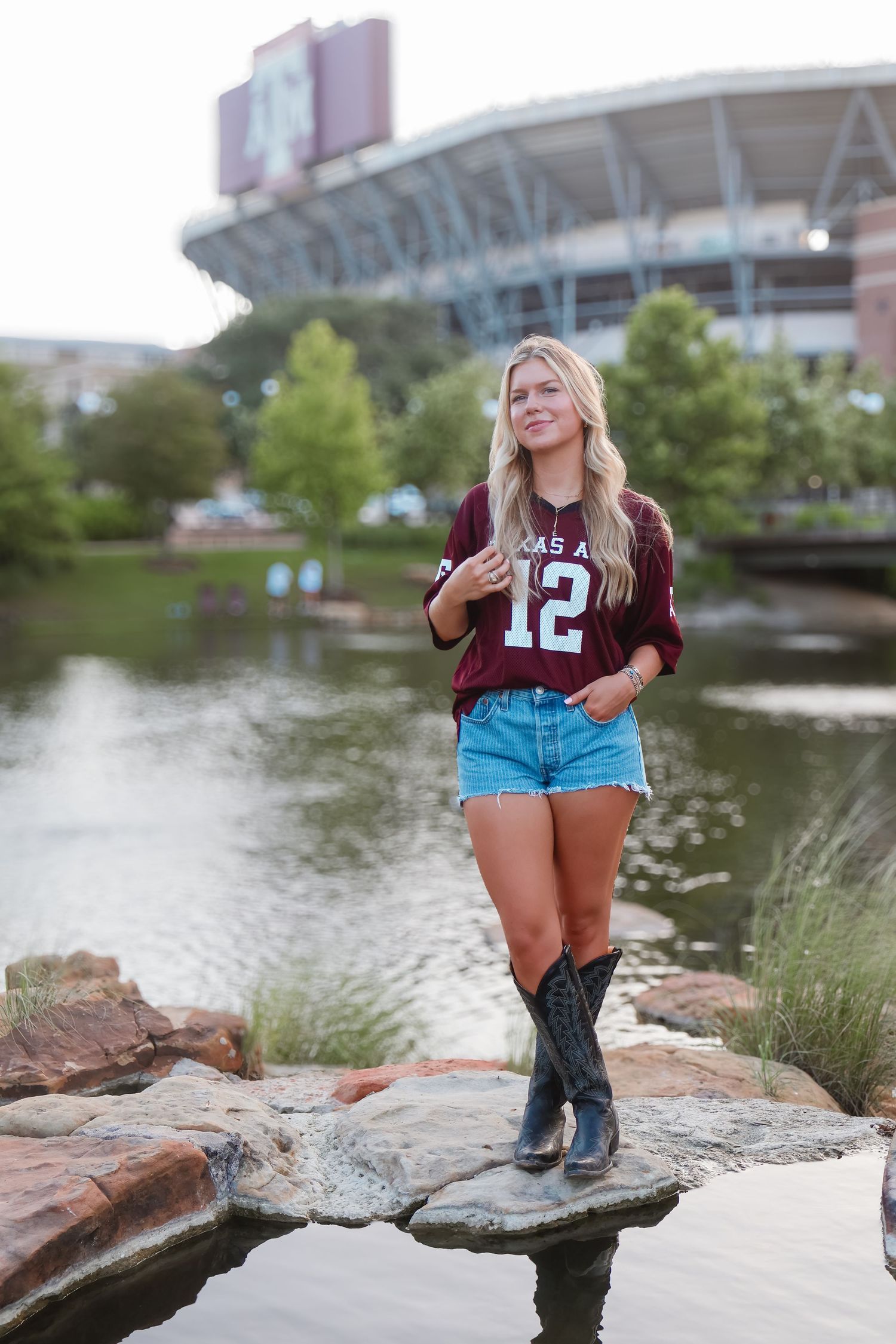 Person in Texas A&M jersey and boots sitting on rocks near a stadium lake.