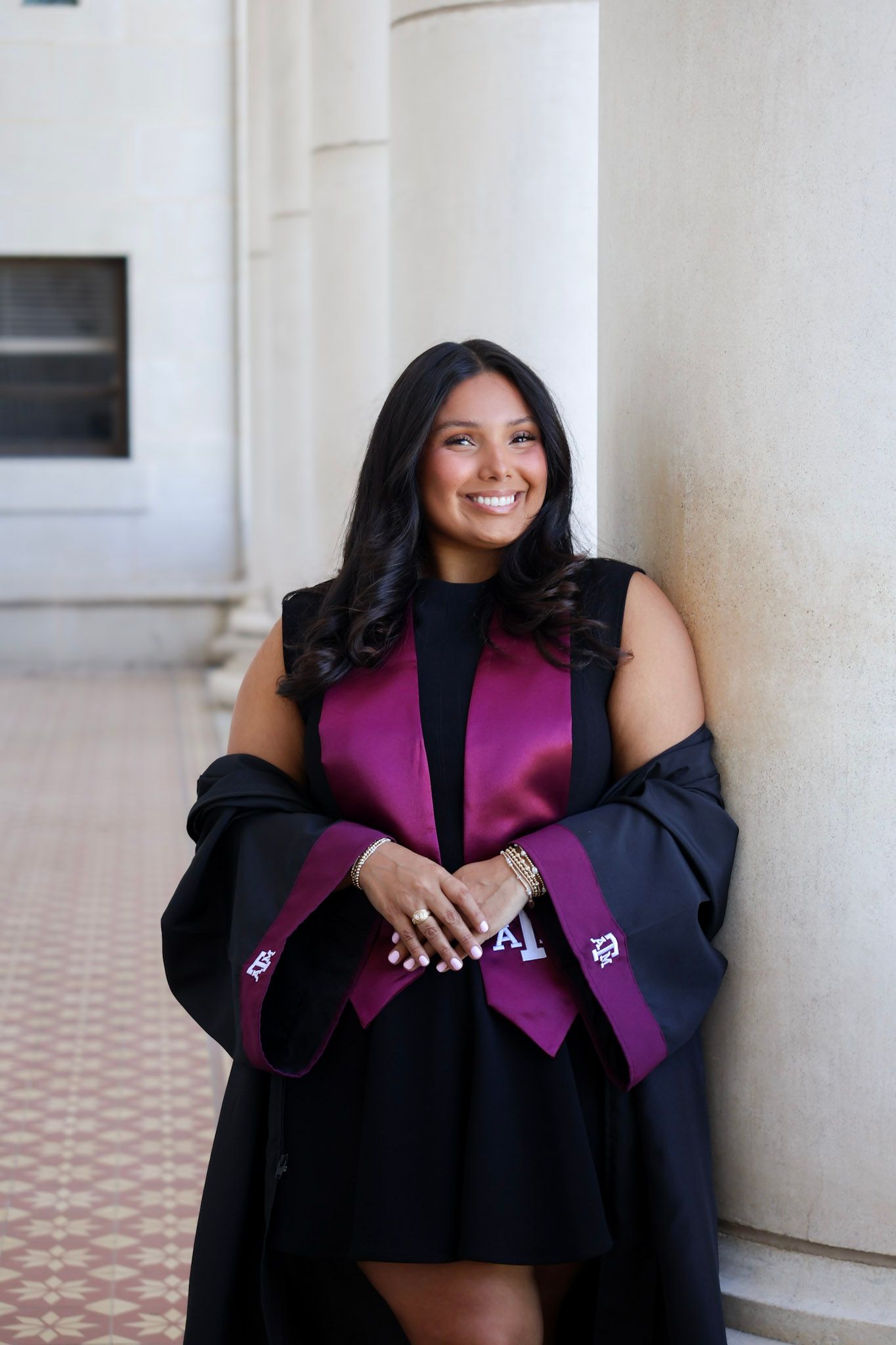 Smiling woman in black graduation gown with maroon stole leaning against column.