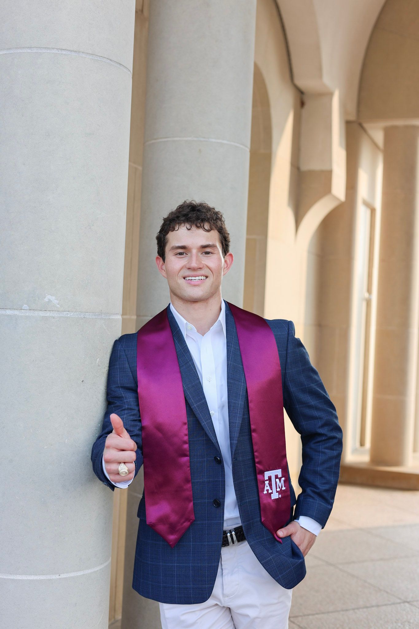 A smiling young man in a navy blazer and maroon Texas A&M graduation stole gives a thumbs up near stone columns.