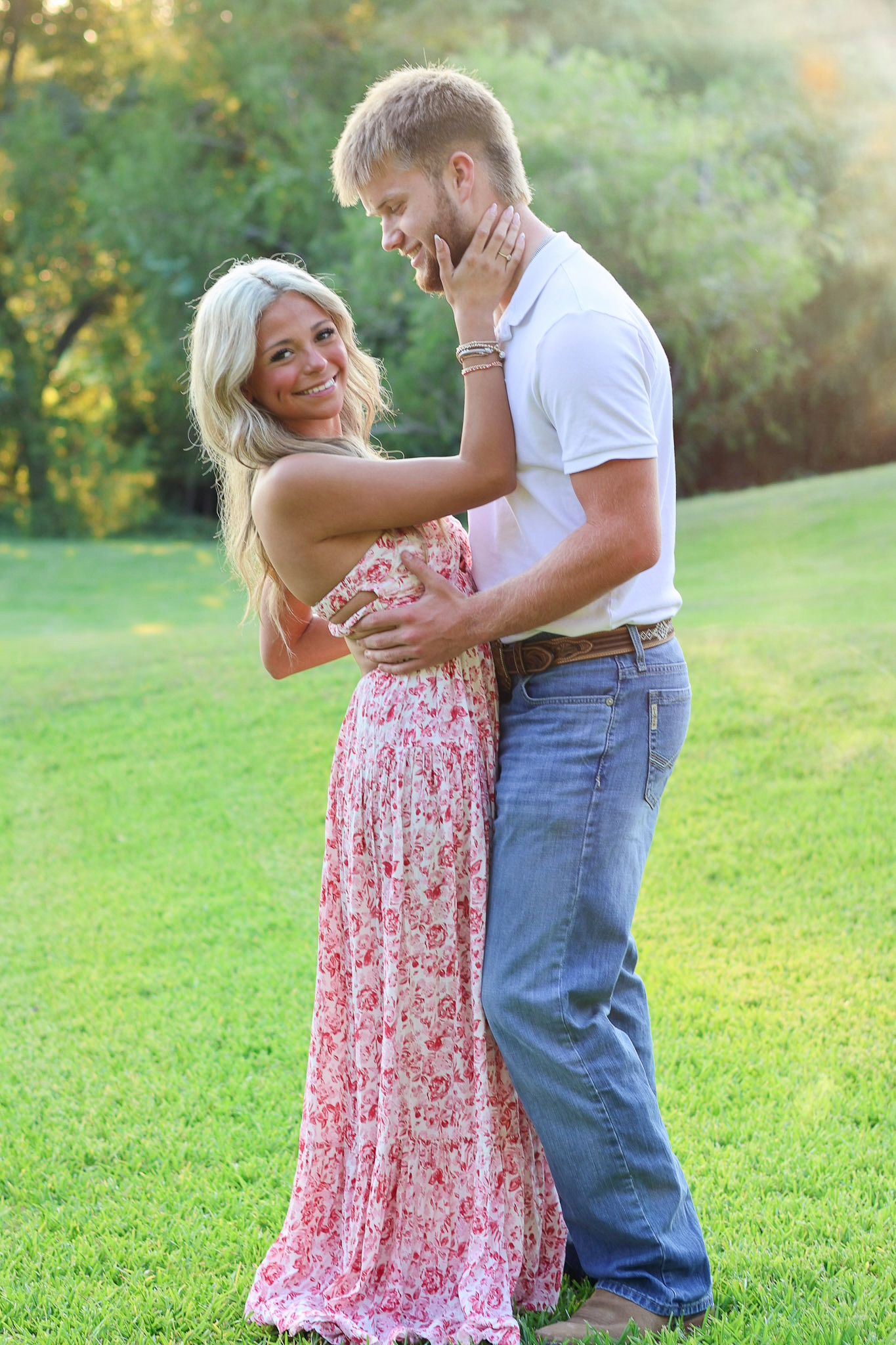 A couple embraces on a grassy hill during a sunny afternoon, with the woman wearing a floral maxi dress.