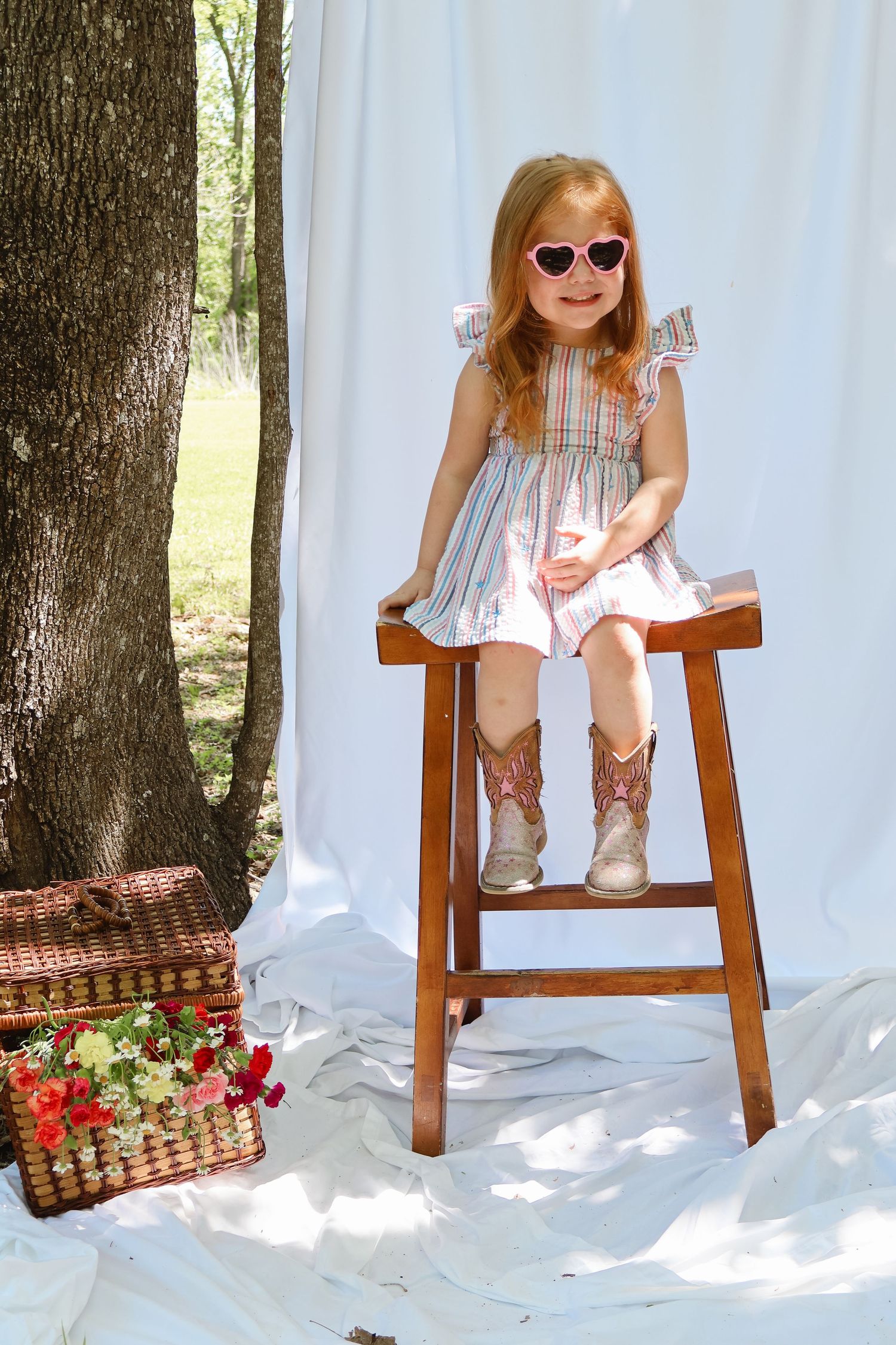 Child sitting on wooden stool wearing pink dress against white backdrop in outdoor photoshoot setting.