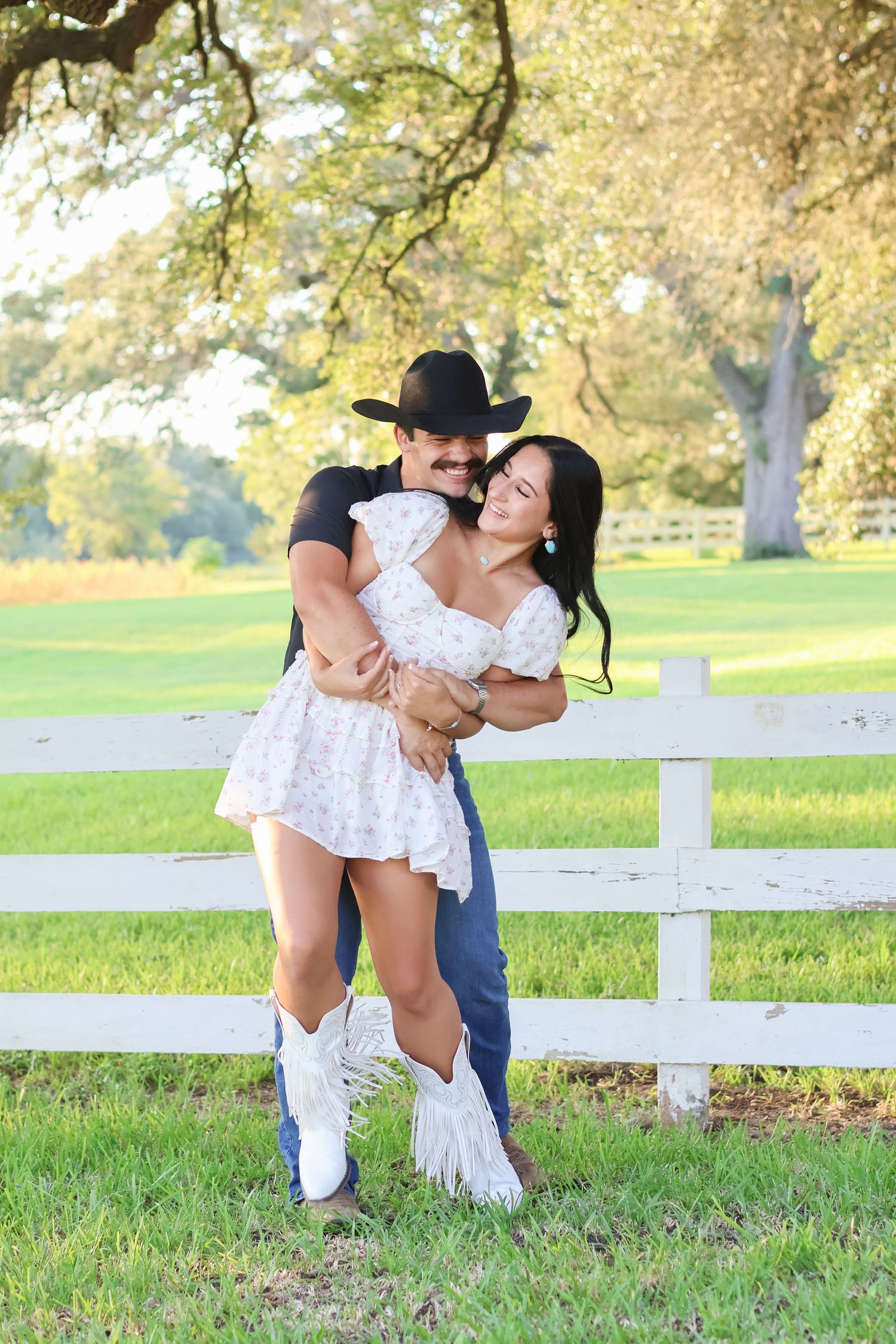 A romantic couple in casual attire embraces playfully on grassy lawn near white fence during golden hour.