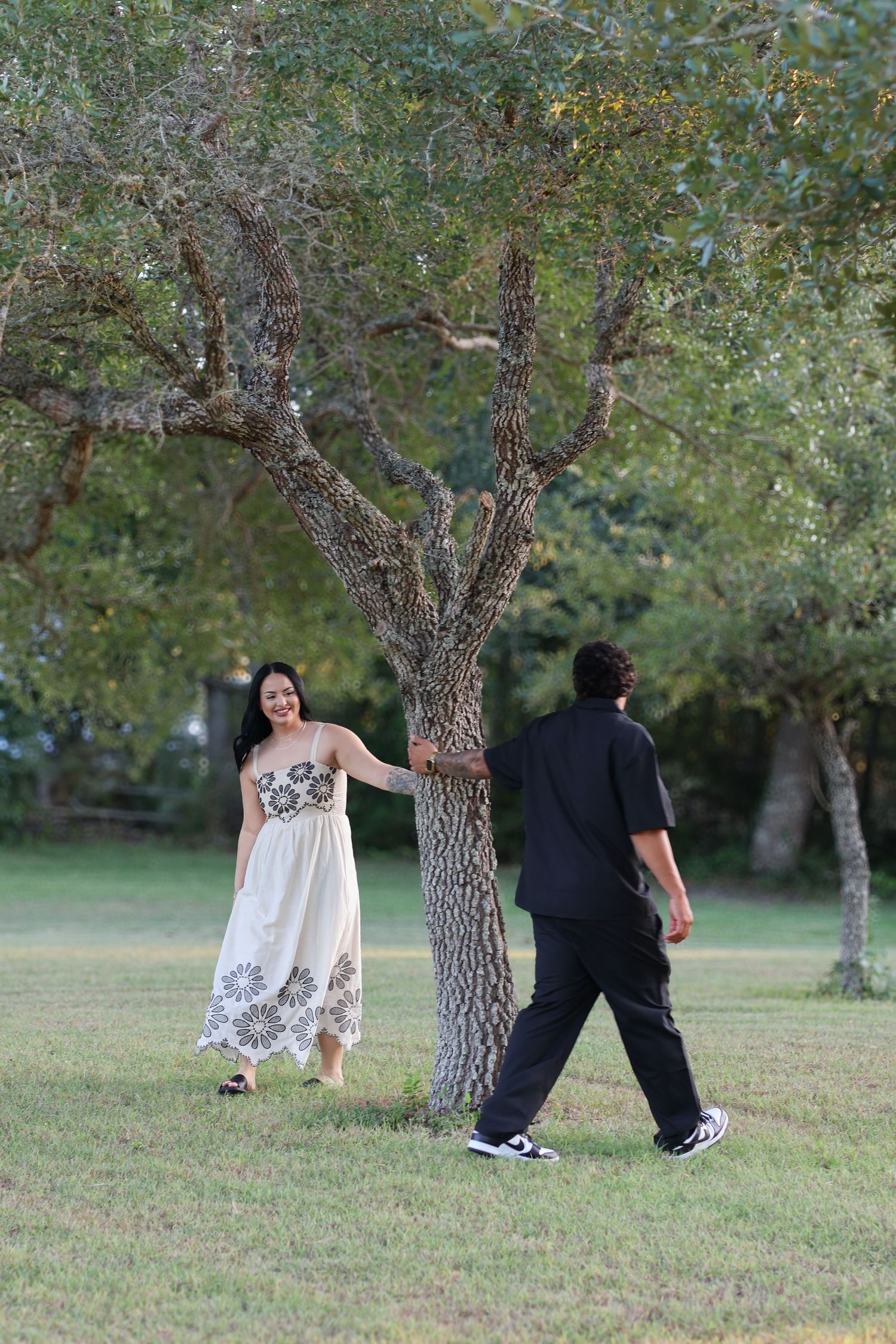 Two people in contrasting black and white outfits walk together through a peaceful park setting with mature trees.