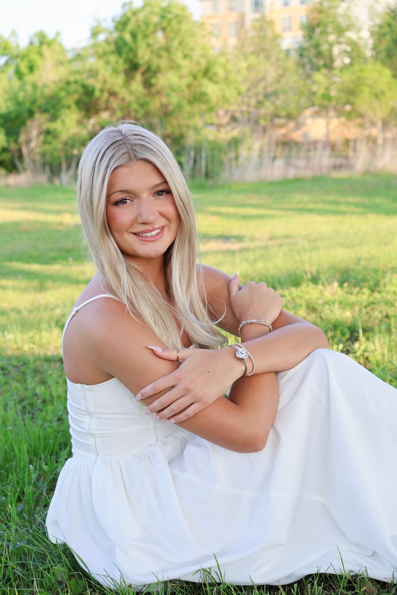 Smiling blonde woman in white dress sitting on green grass outdoors.