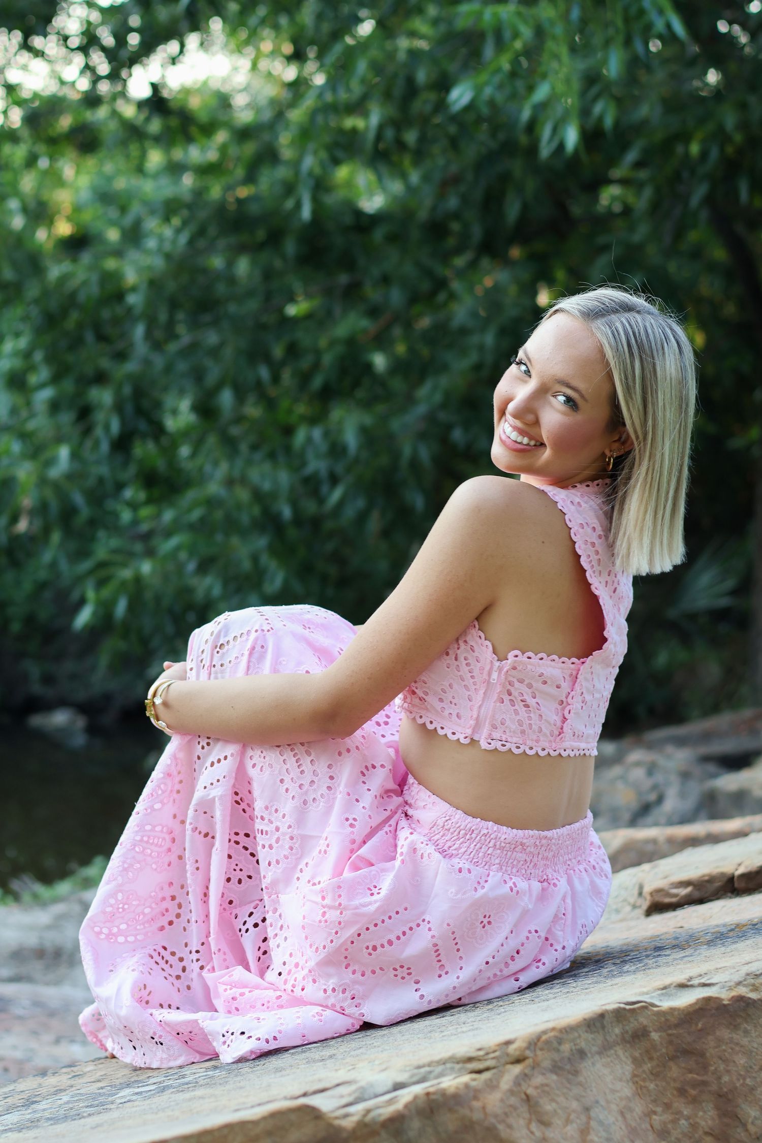 A person in a pink eyelet dress poses gracefully on a stone ledge in a natural outdoor setting.