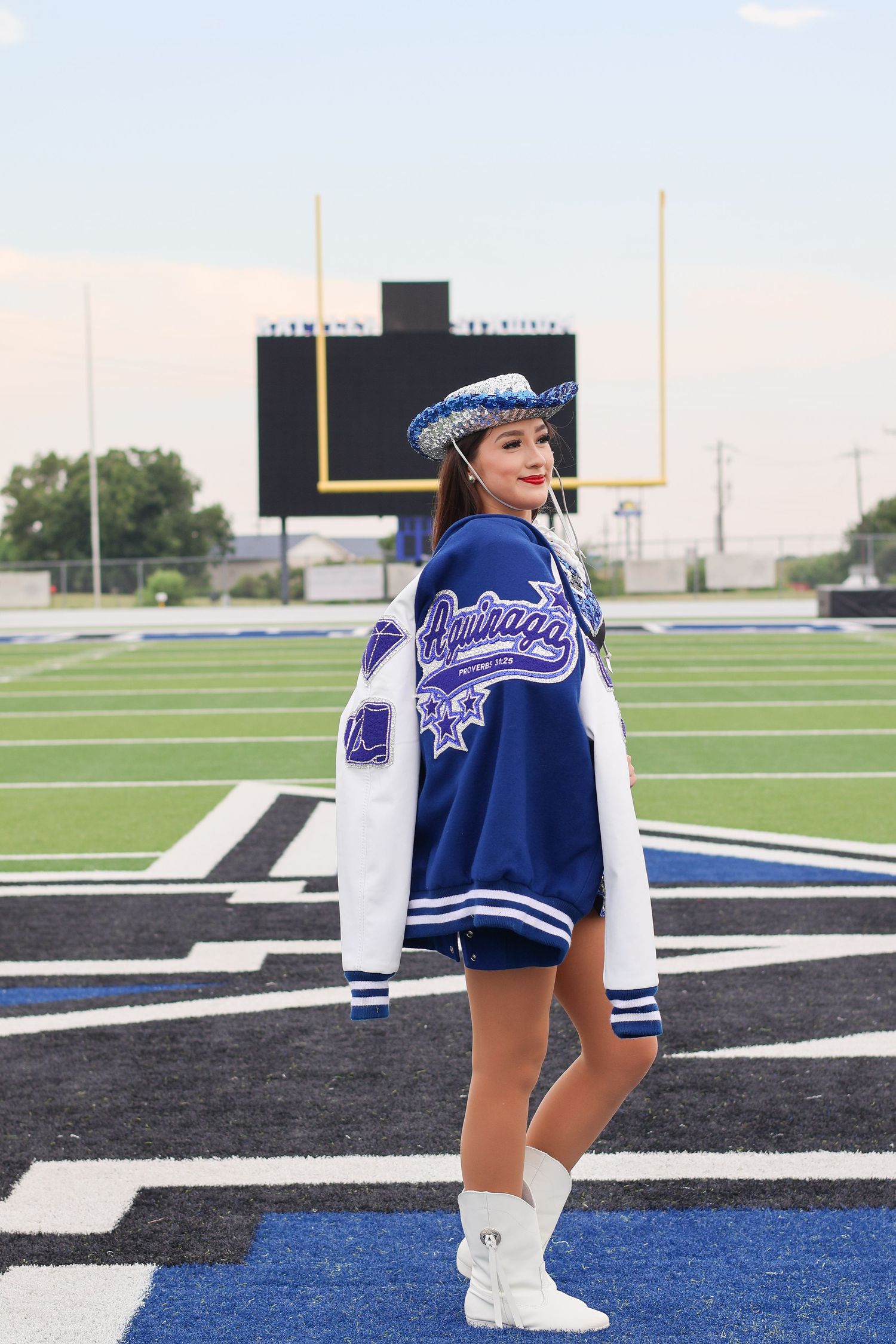 Cheerleader in blue and white varsity jacket poses on football field goal line with scoreboard in background.