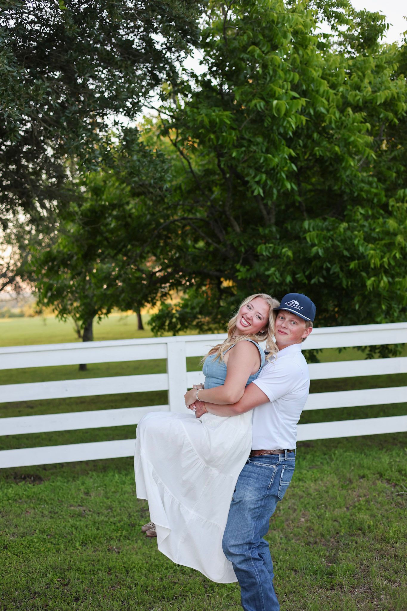 A man in jeans and a cap joyfully lifts a smiling woman in a white skirt near a white fence outdoors.