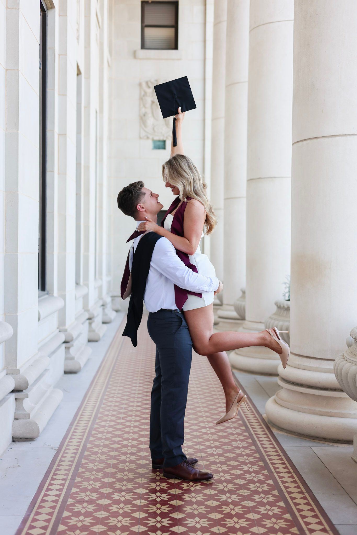 Couple celebrating graduation in elegant columned hallway, man lifting woman holding mortarboard cap, wearing formal attire.