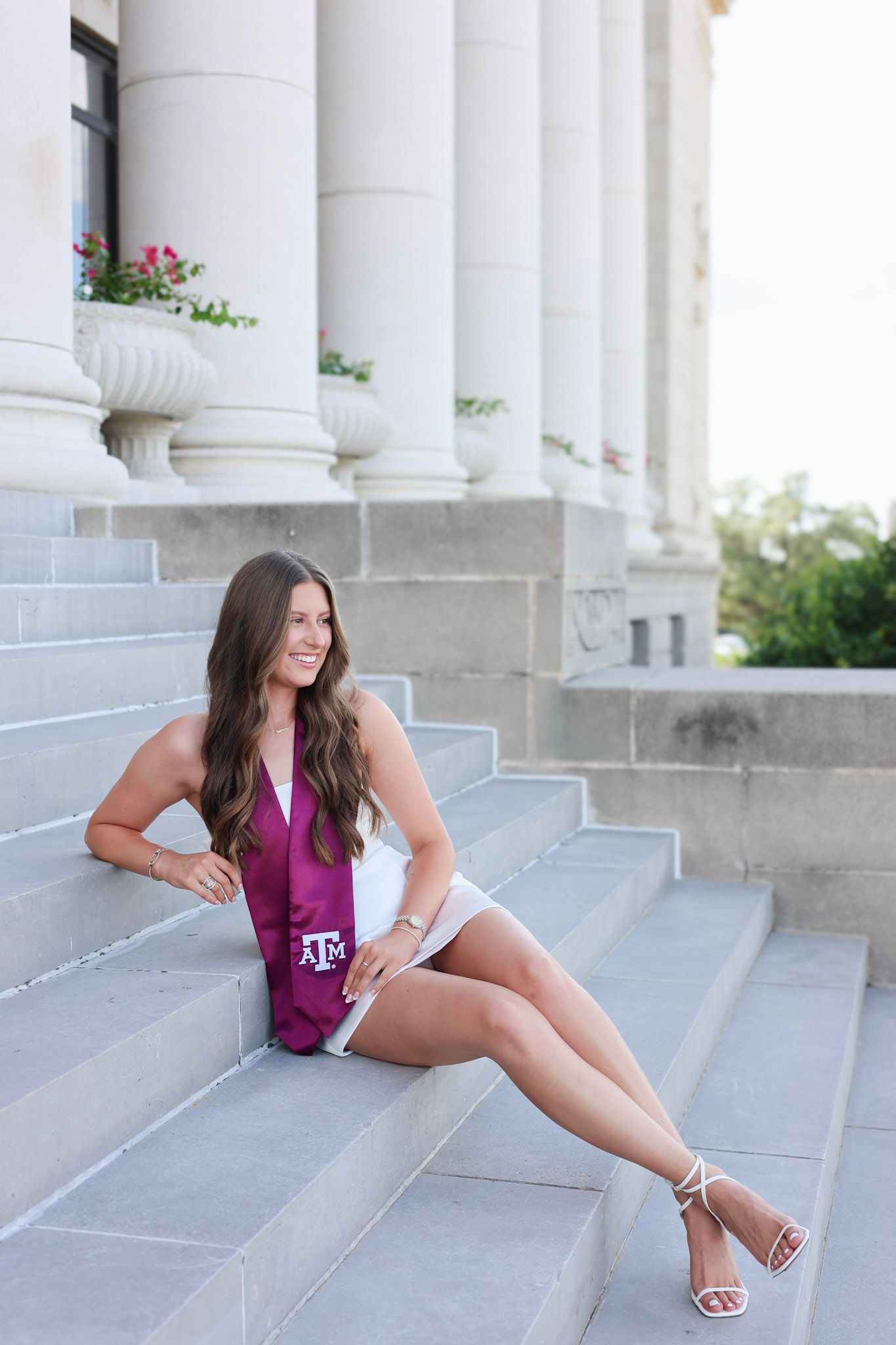 Brunette woman in white shorts with maroon Texas A&M graduation stole sitting on steps by white columns.