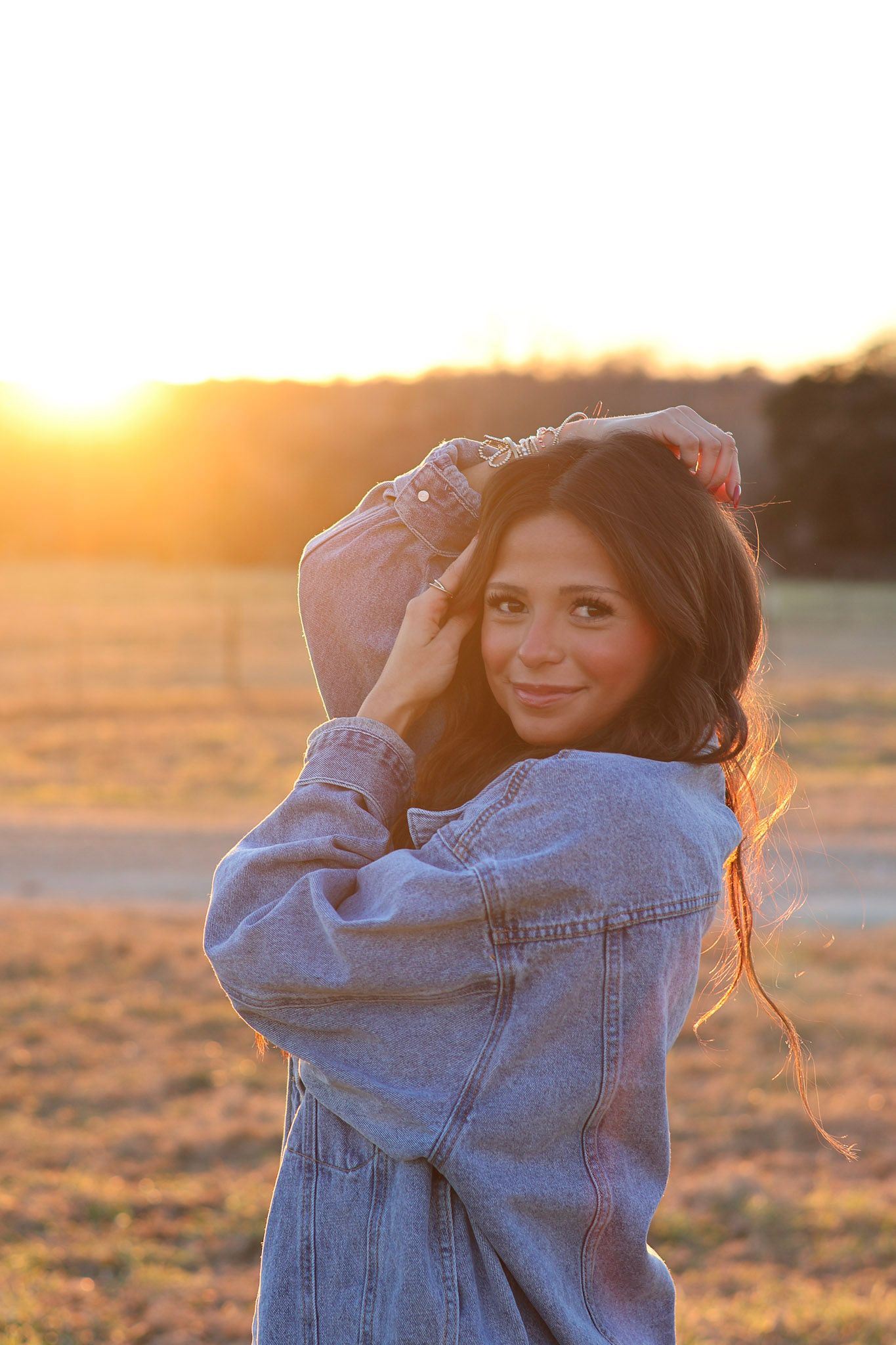 Brunette woman in oversized denim jacket posing in sunlit field at golden hour sunset.