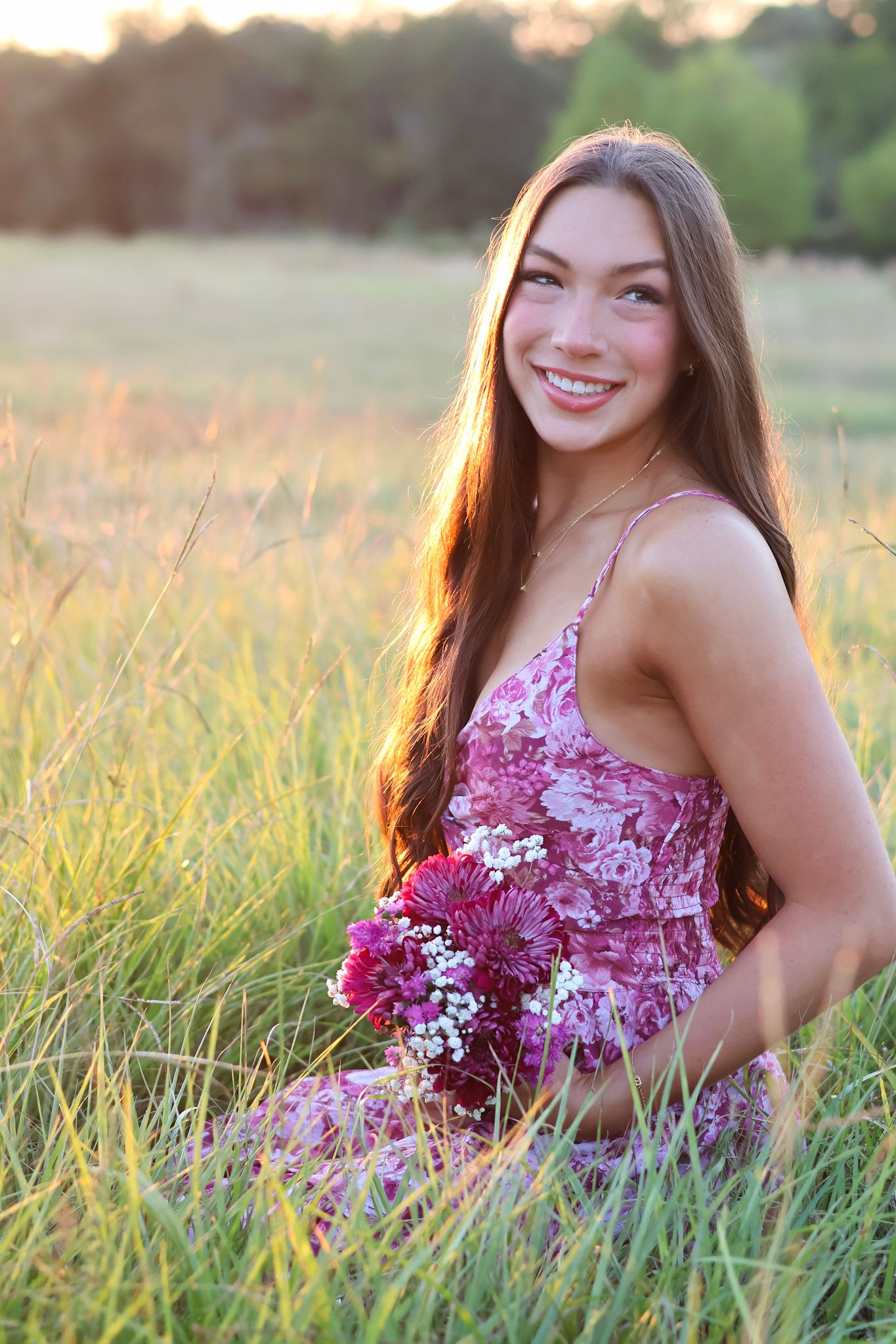 Smiling young woman in floral dress sitting in golden field holding purple flowers at sunset.