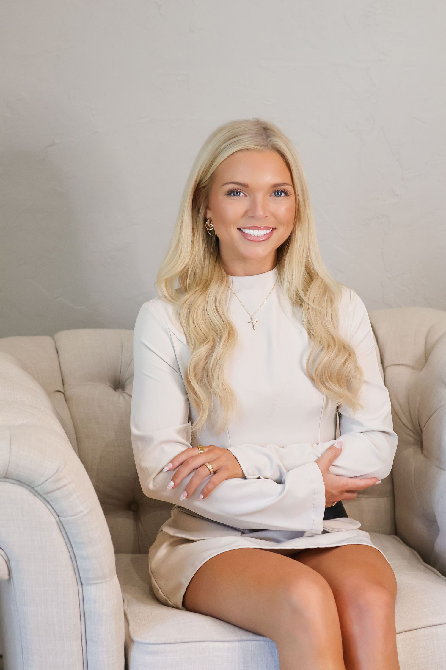 Person in white sweater smiling and posing on light gray couch in modern setting.