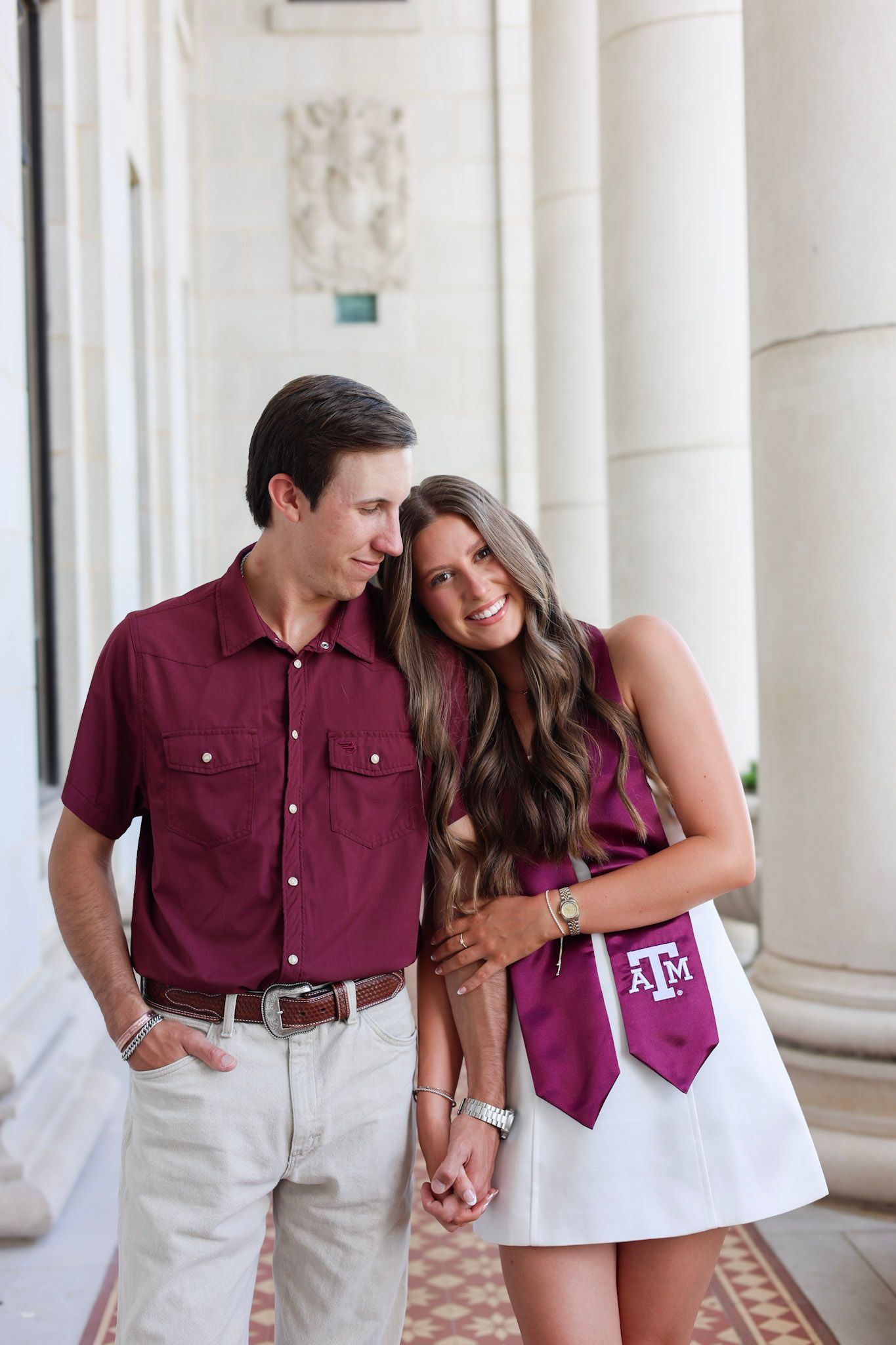 Texas A&M graduate in maroon stole and white dress poses with partner in matching maroon shirt between columns.