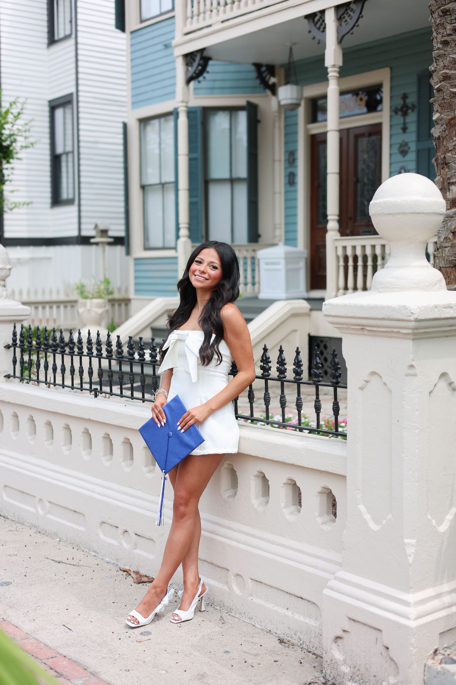Young woman in white dress holding blue graduation cap poses by ornate white fence near Victorian home.