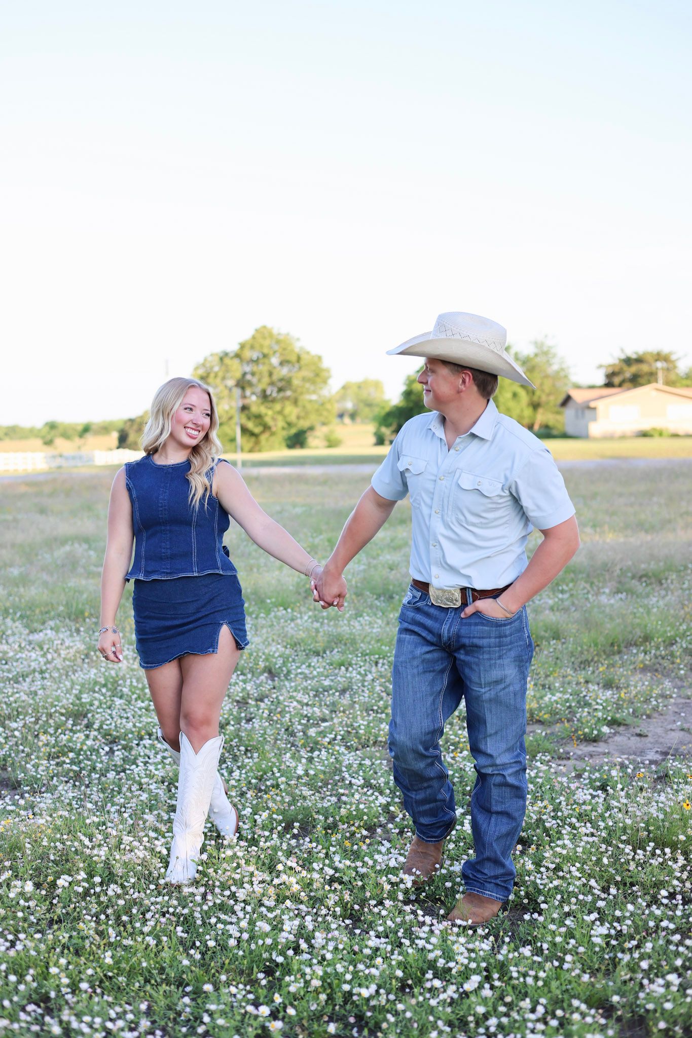 A couple holds hands in a wildflower field, woman in blue dress and white boots, man in cowboy hat and jeans.