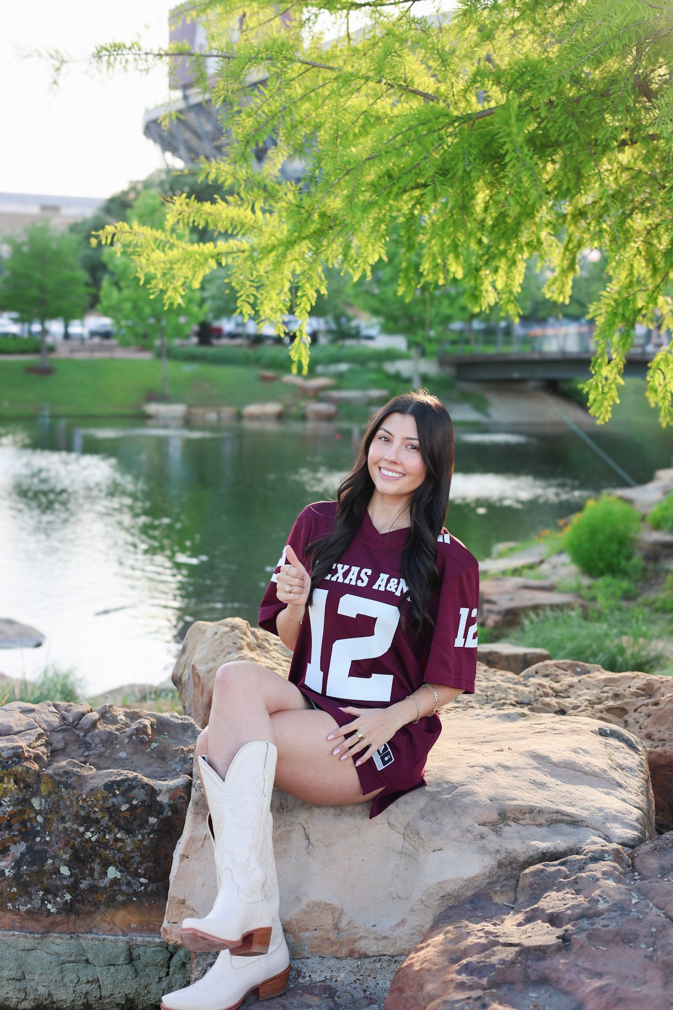 Young woman in Texas A&M jersey and white cowboy boots sitting on rocks by a pond.