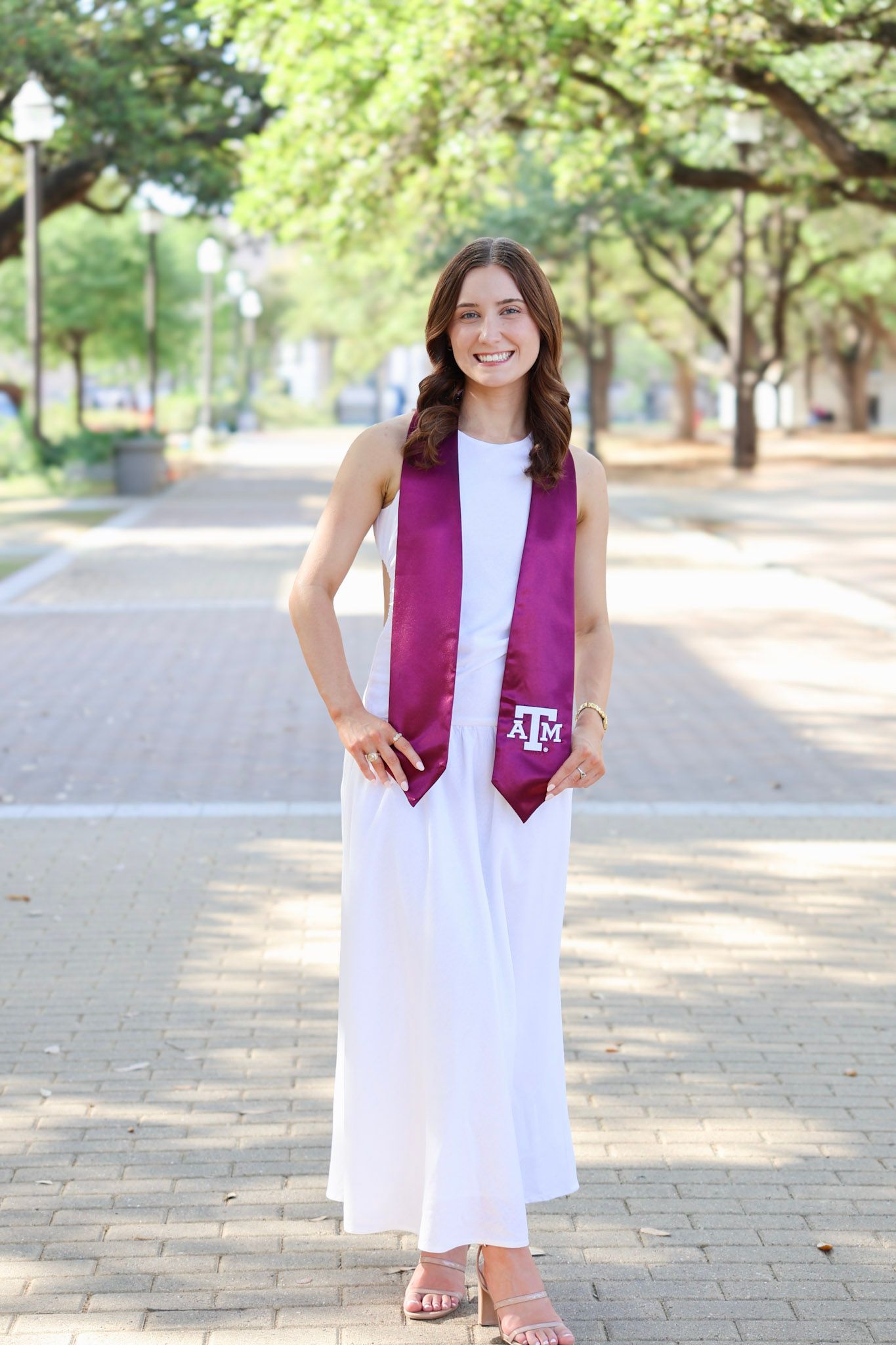 Smiling young woman in white dress wearing Texas A&M maroon graduation stole poses on a tree-lined campus walkway.
