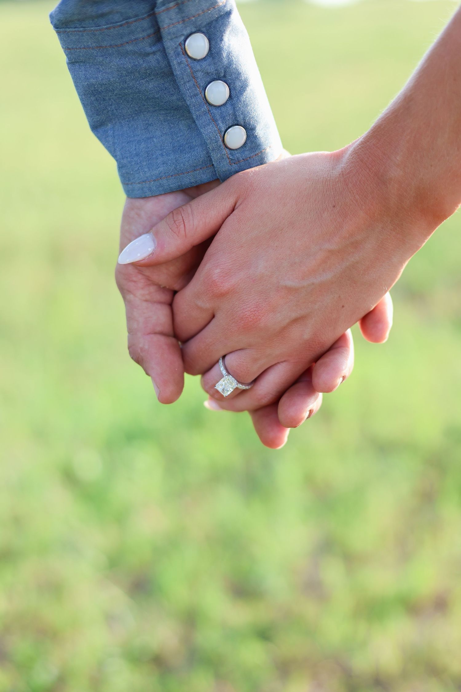 Close-up of couple holding hands outdoors showing diamond engagement ring, green grass background.