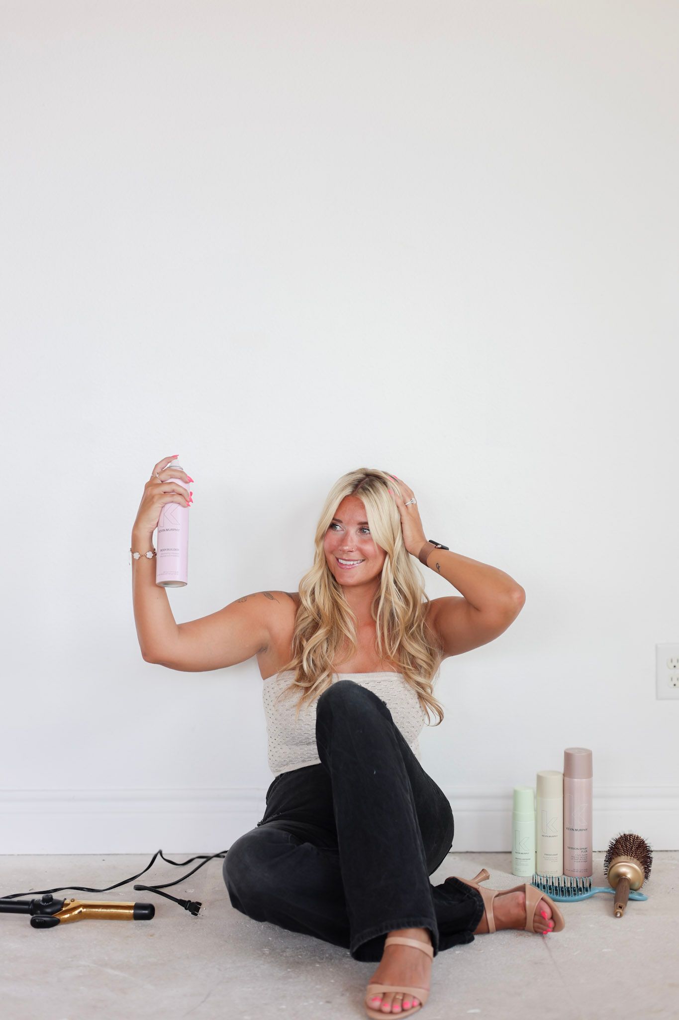 Blonde woman sitting on floor holding pink hair spray, surrounded by styling tools and hair products against white wall.