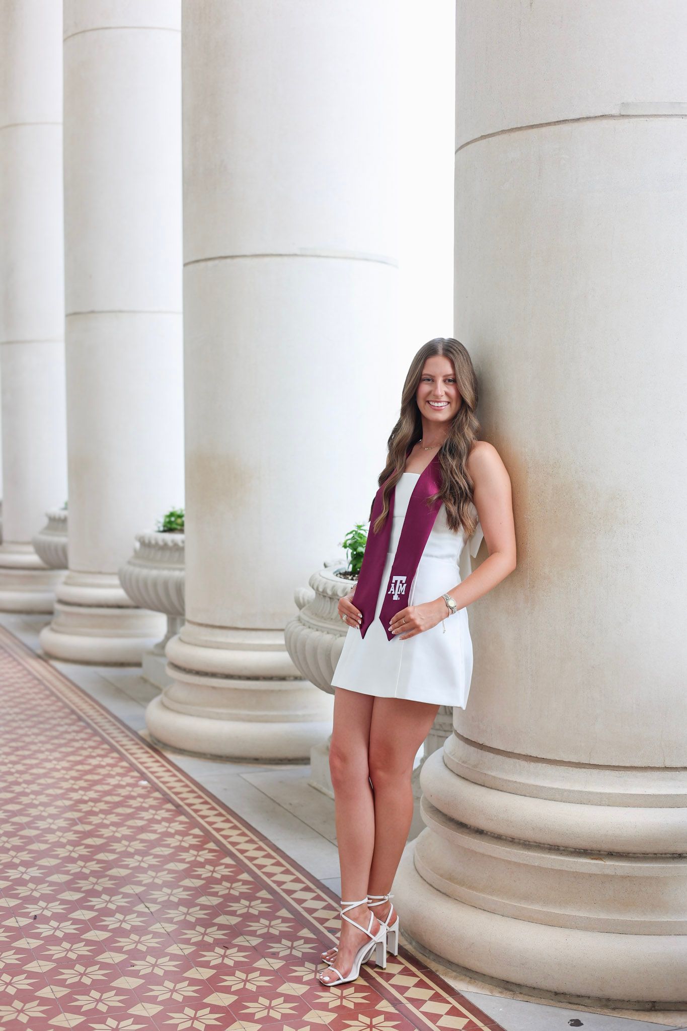 Young woman in white dress with maroon Texas A&M graduation stole posing between white columns.