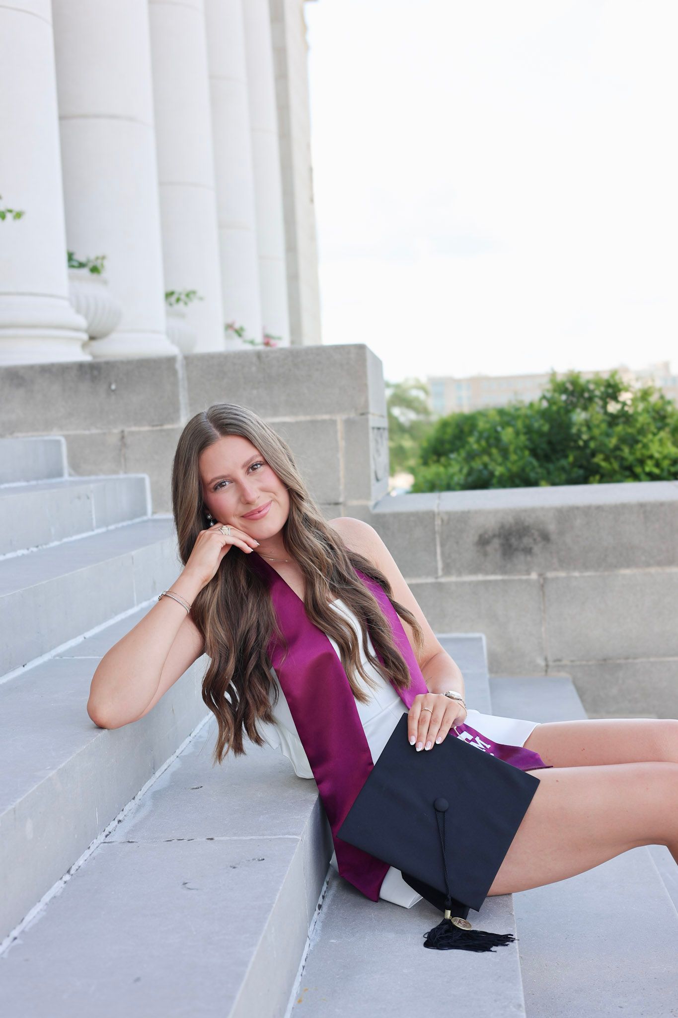 Brunette graduate posing on stone steps with purple sash and mortarboard cap outdoors.