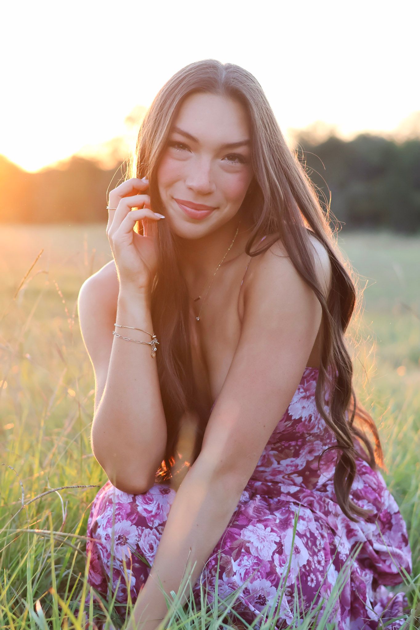 Young woman in floral dress sitting in golden hour sunlight in a grassy field, smiling softly at camera.