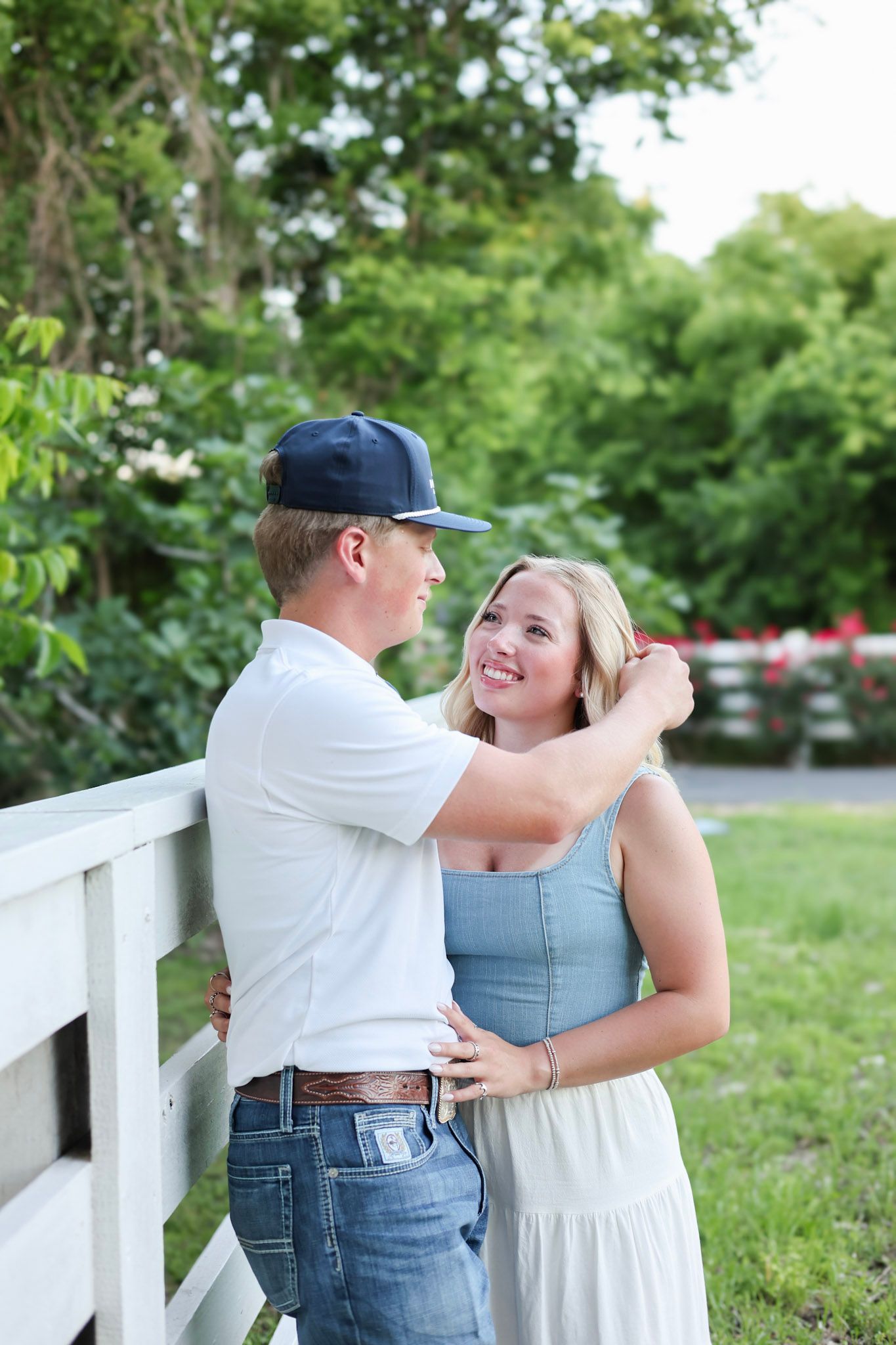 A young couple shares a romantic moment by a white fence, surrounded by lush greenery, as he tenderly touches her hair.