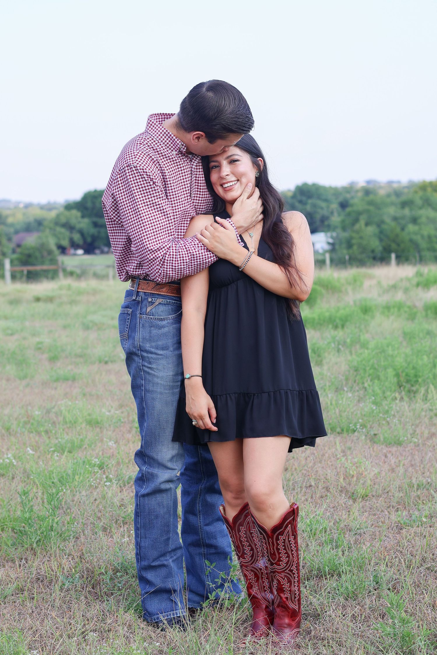 A couple embraces in a grassy field while wearing western-inspired outfits with cowboy boots and plaid shirt.