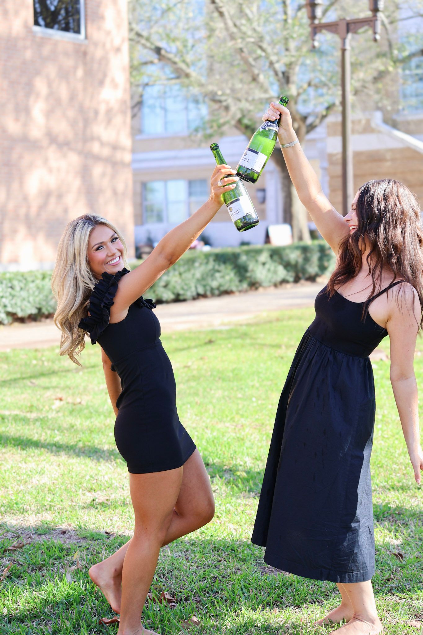Two women in black dresses toasting with green wine bottles on a sunny lawn.