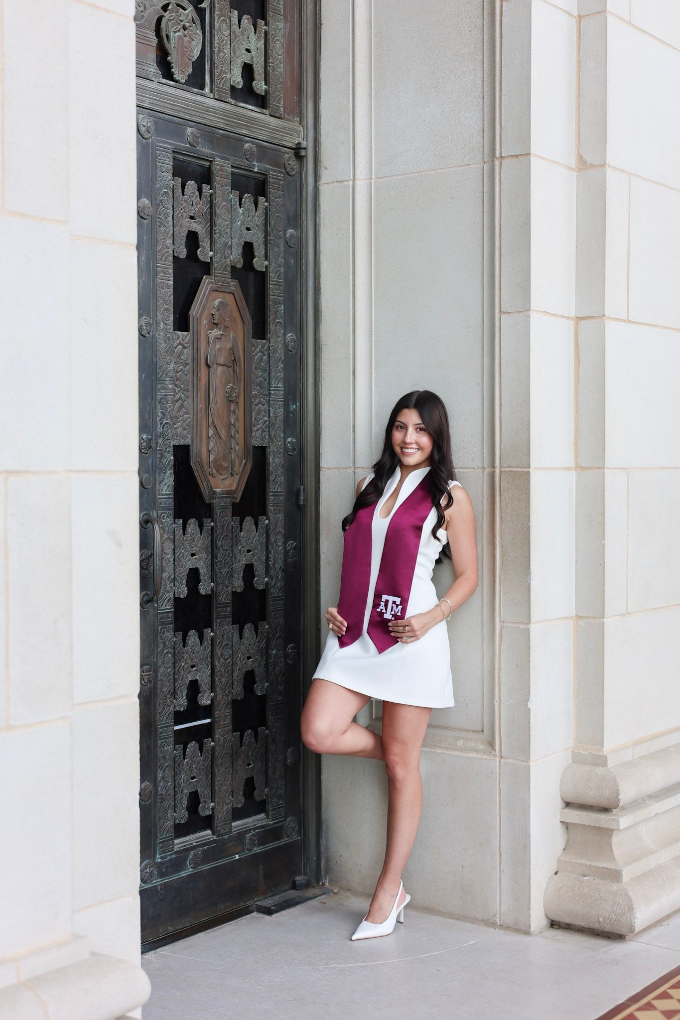 Texas A&M graduate poses in white dress with maroon Aggie sash by ornate campus building doors.