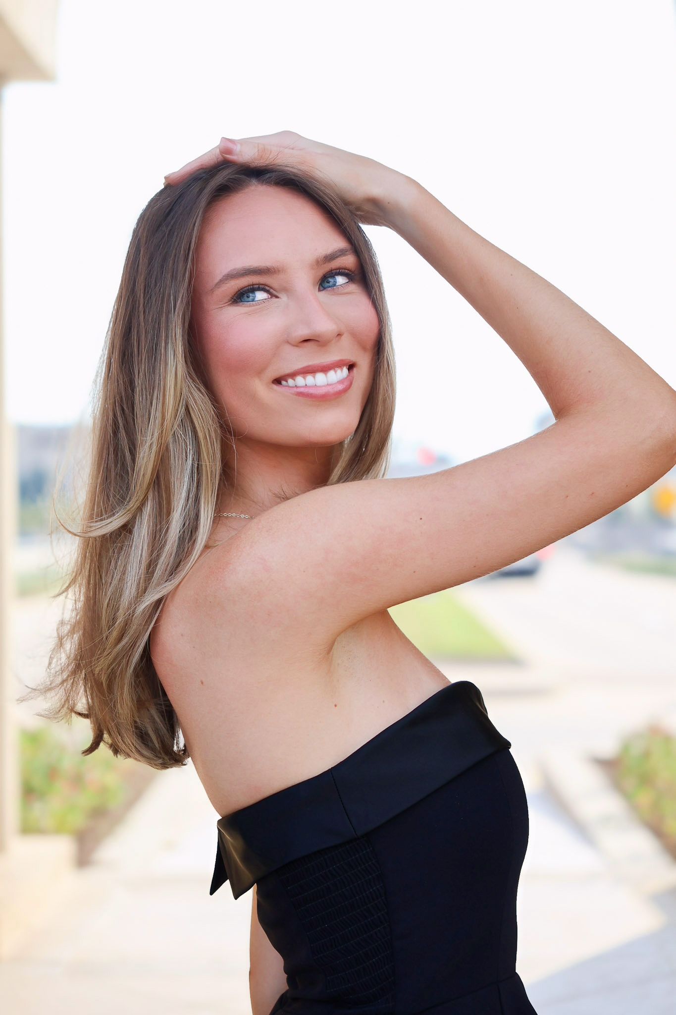 Woman in black strapless dress smiles while running hand through long blonde hair outdoors.