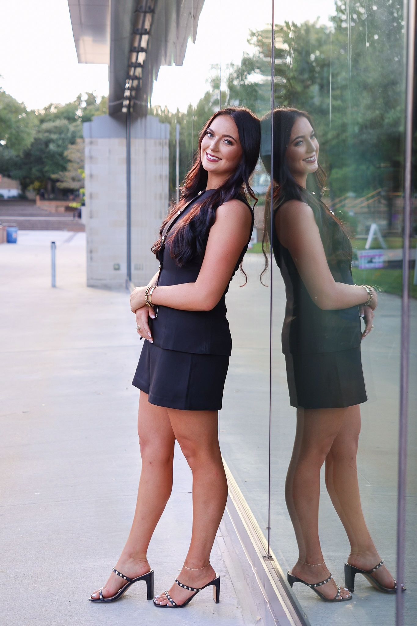 Woman in black dress and heels poses against reflective glass window at sunset.