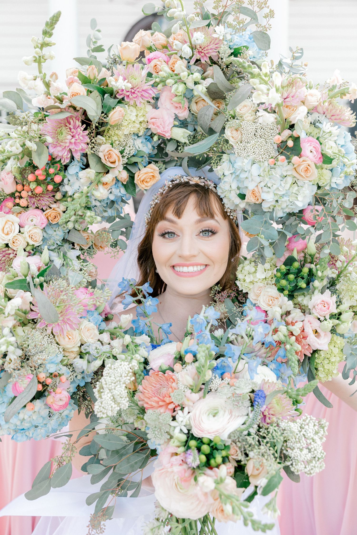 Smiling bride surrounded by lush pastel floral arch with pink, blue, and white blooms, holding a matching bouquet.