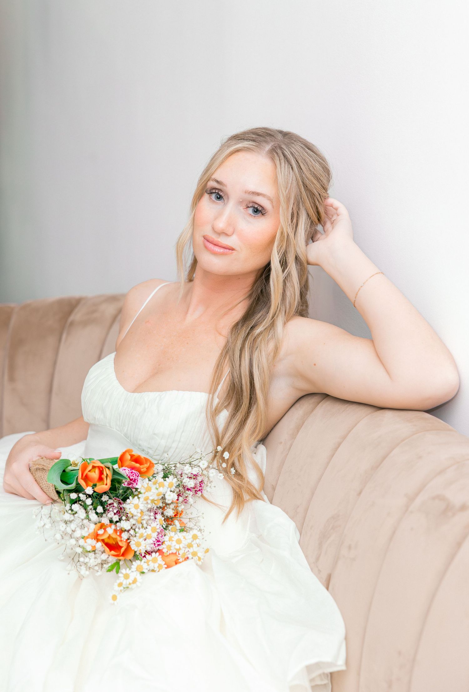 Blonde bride in white gown sits on beige sofa holding orange and white bouquet, posing elegantly for bridal portrait.