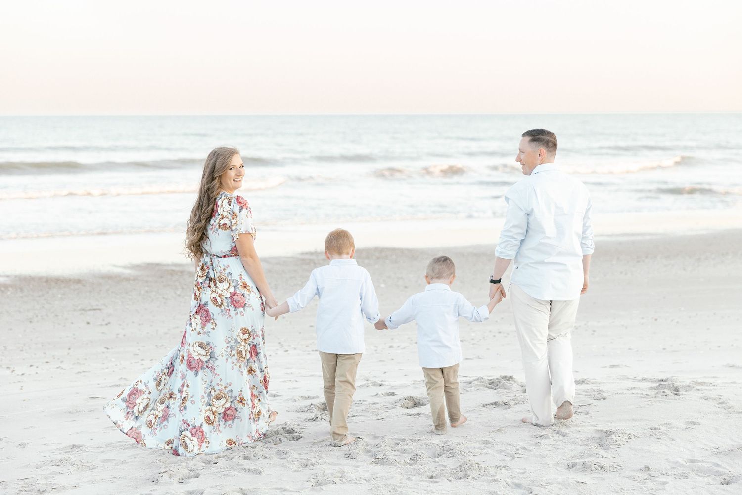 Family of four holding hands walking toward ocean waves on a sandy beach at sunset in white and beige outfits.