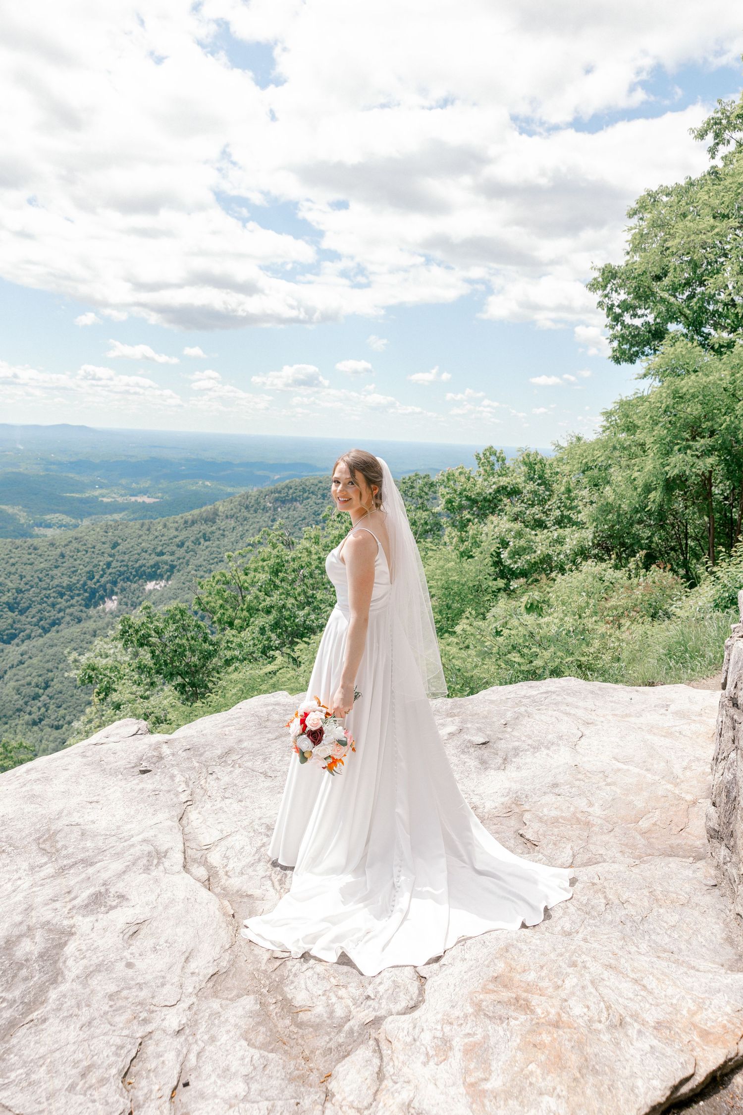 A bride in white dress poses on a rocky mountain overlook with scenic valley views.