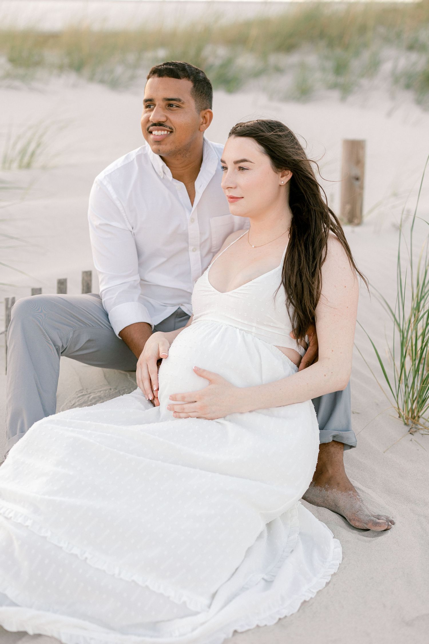 Expectant couple sits on beach in white attire for maternity photoshoot.
