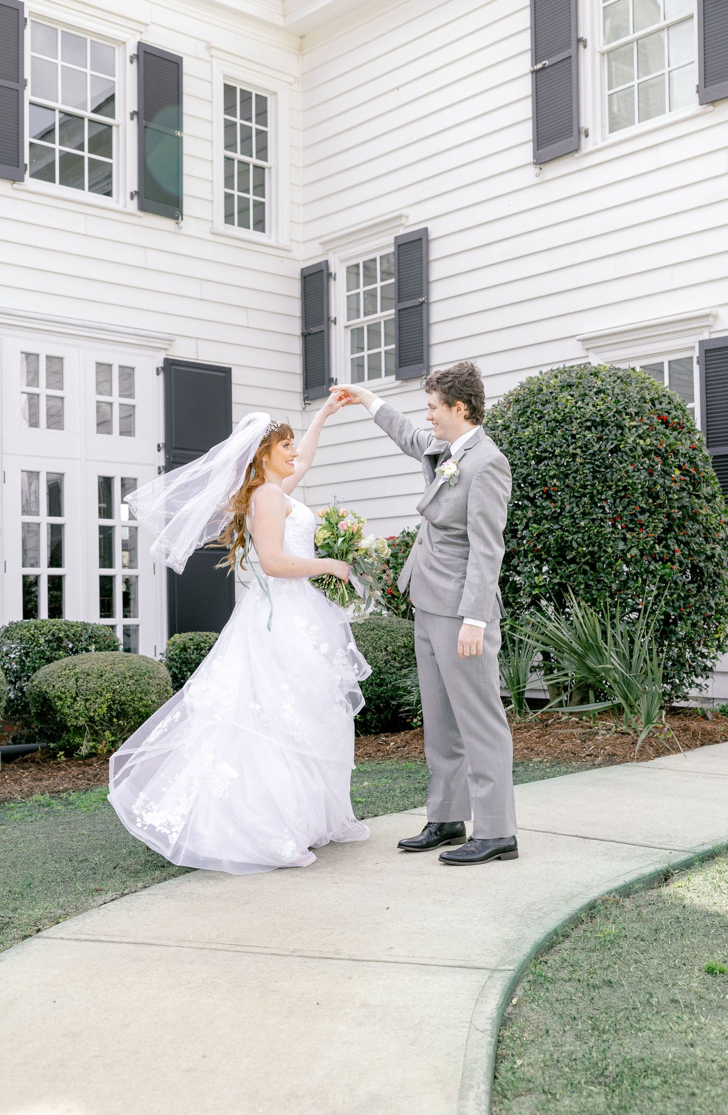 Bride and groom dancing outside white colonial house with black shutters.