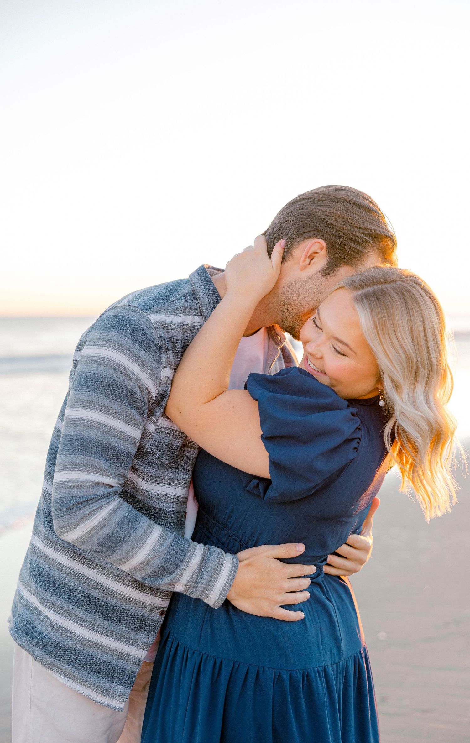 Romantic couple embracing on beach at sunset wearing blue and striped outfits.