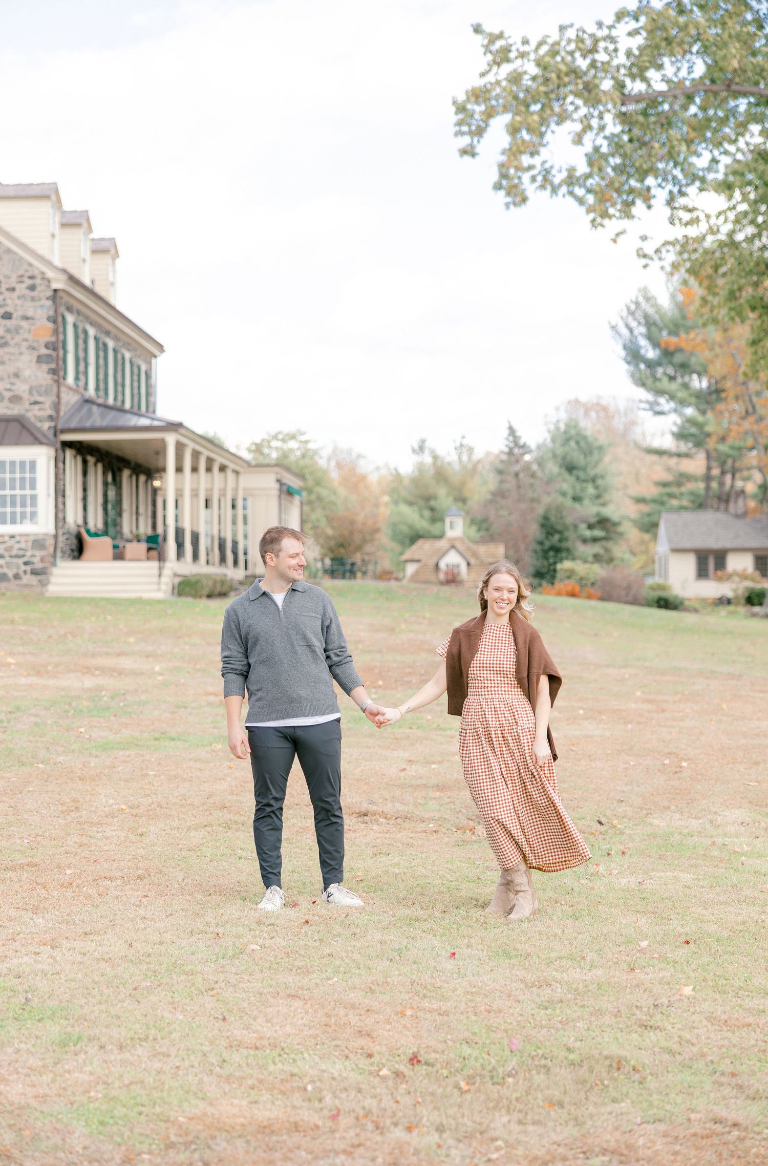 A couple holds hands and smiles while walking on a grassy lawn with a stone estate and autumn trees in the background.