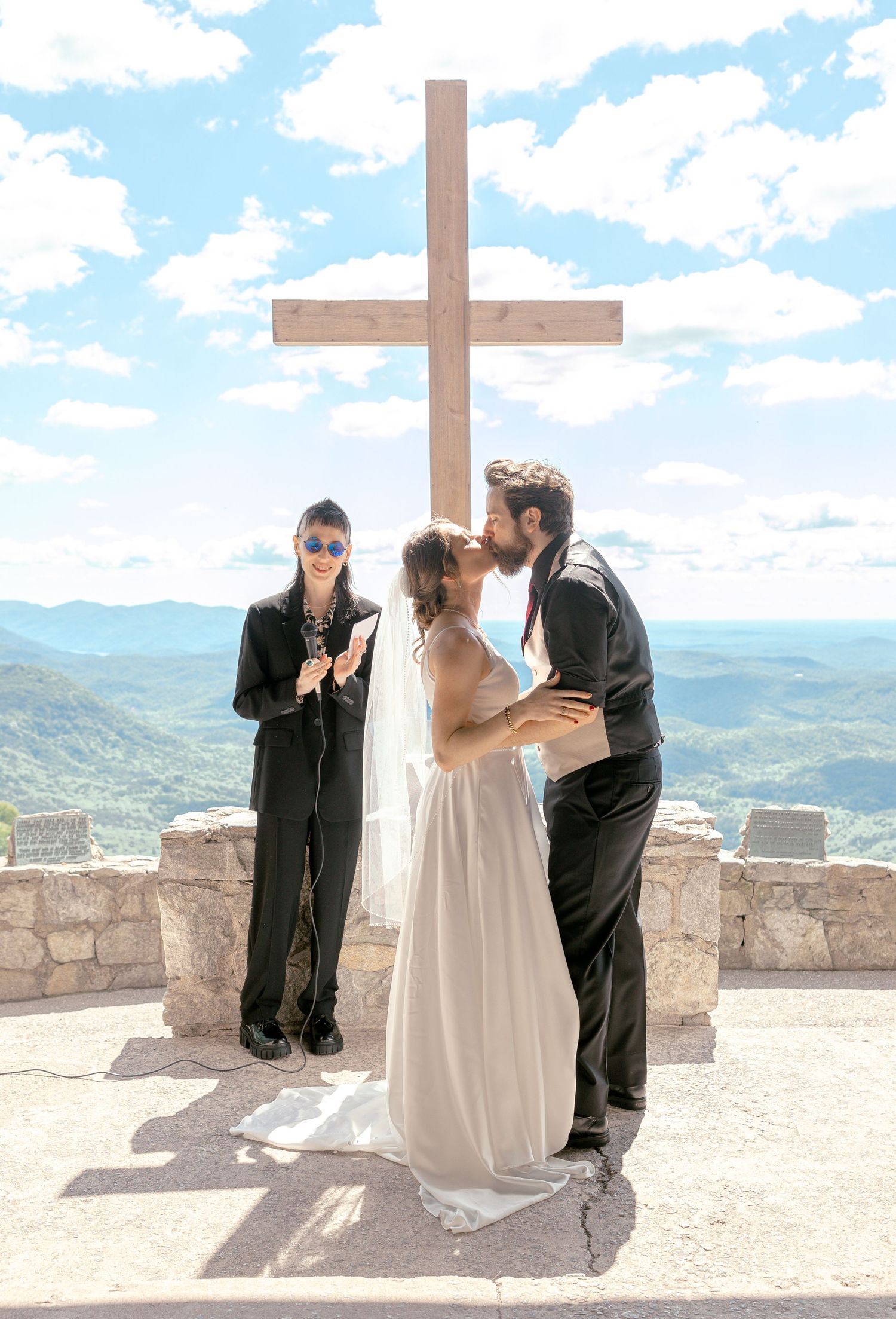 Bride and groom share a kiss at outdoor mountain wedding ceremony before a wooden cross under blue skies.